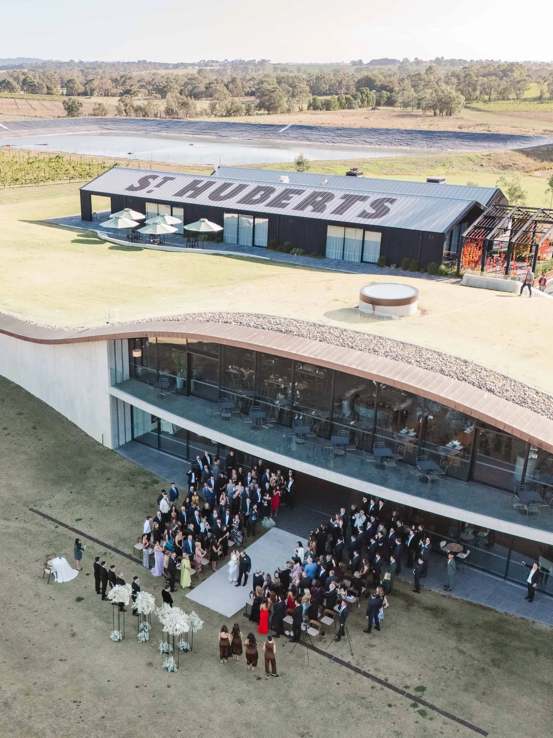 Aerial Melbourne wedding photography of an outdoor ceremony at St Hubert’s, with guests gathered beside modern vineyard architecture.