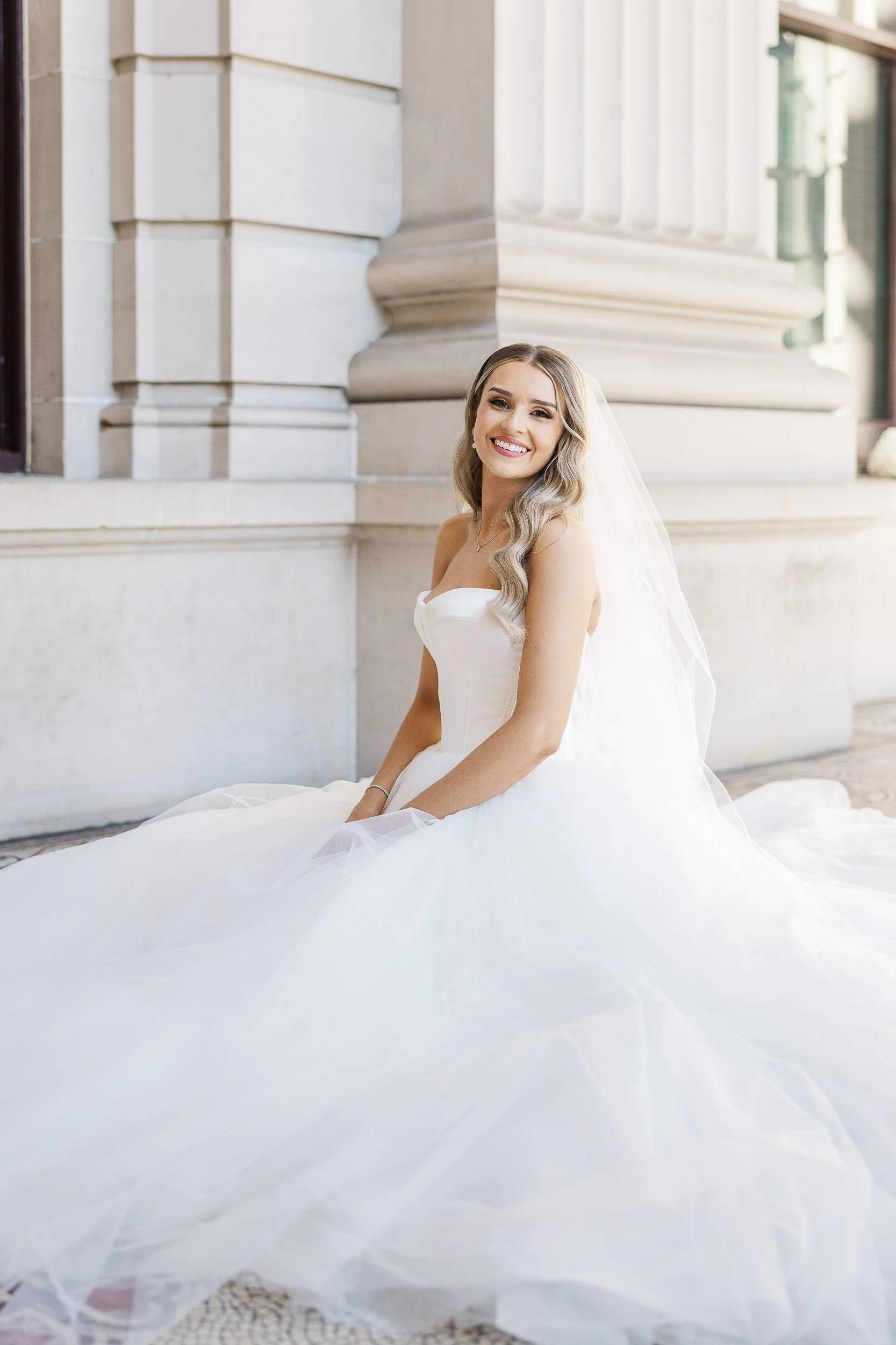 Melbourne wedding photography portrait of a smiling bride in a strapless gown and veil seated beside elegant stone columns of Victoria Parliament House