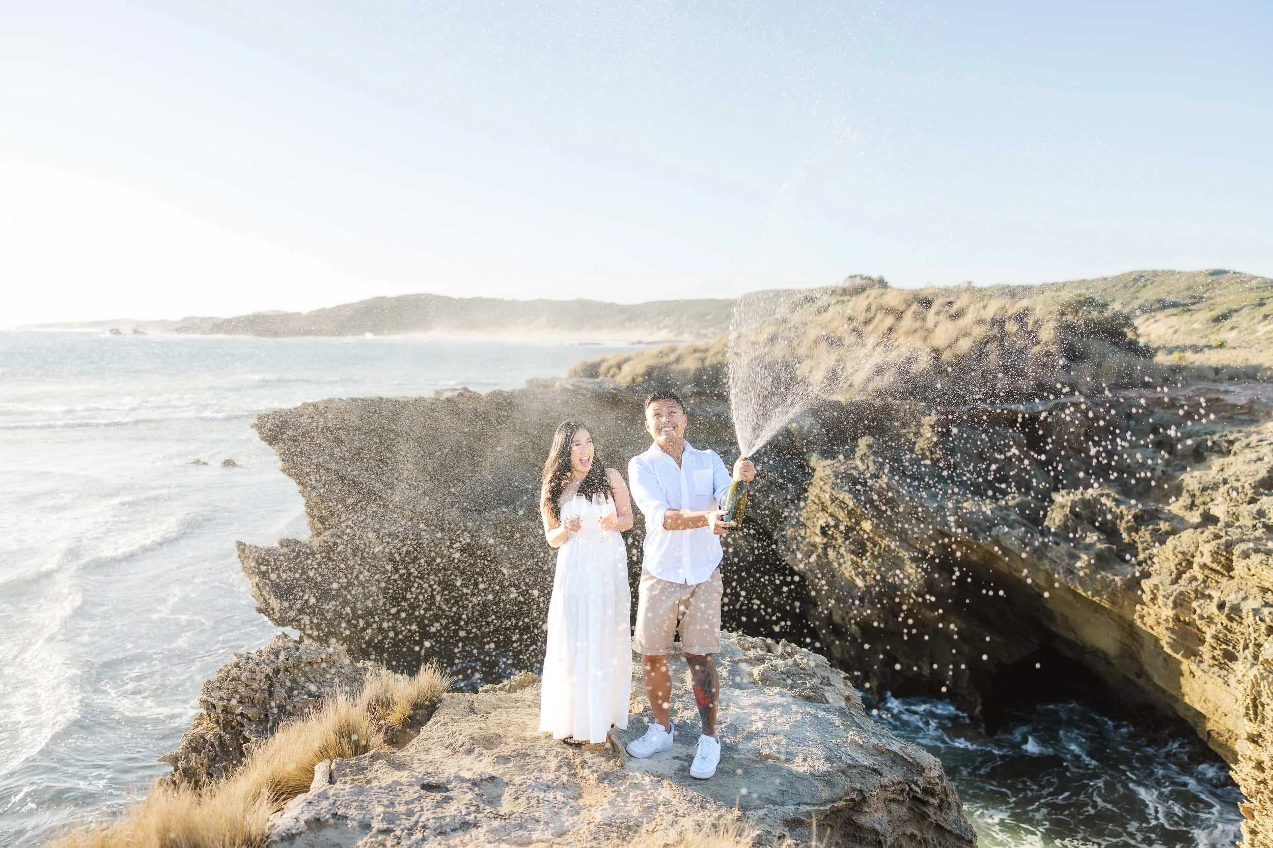 Melbourne Proposal Photography of a couple celebrating on coastal cliffs with champagne spray after a beach proposal.