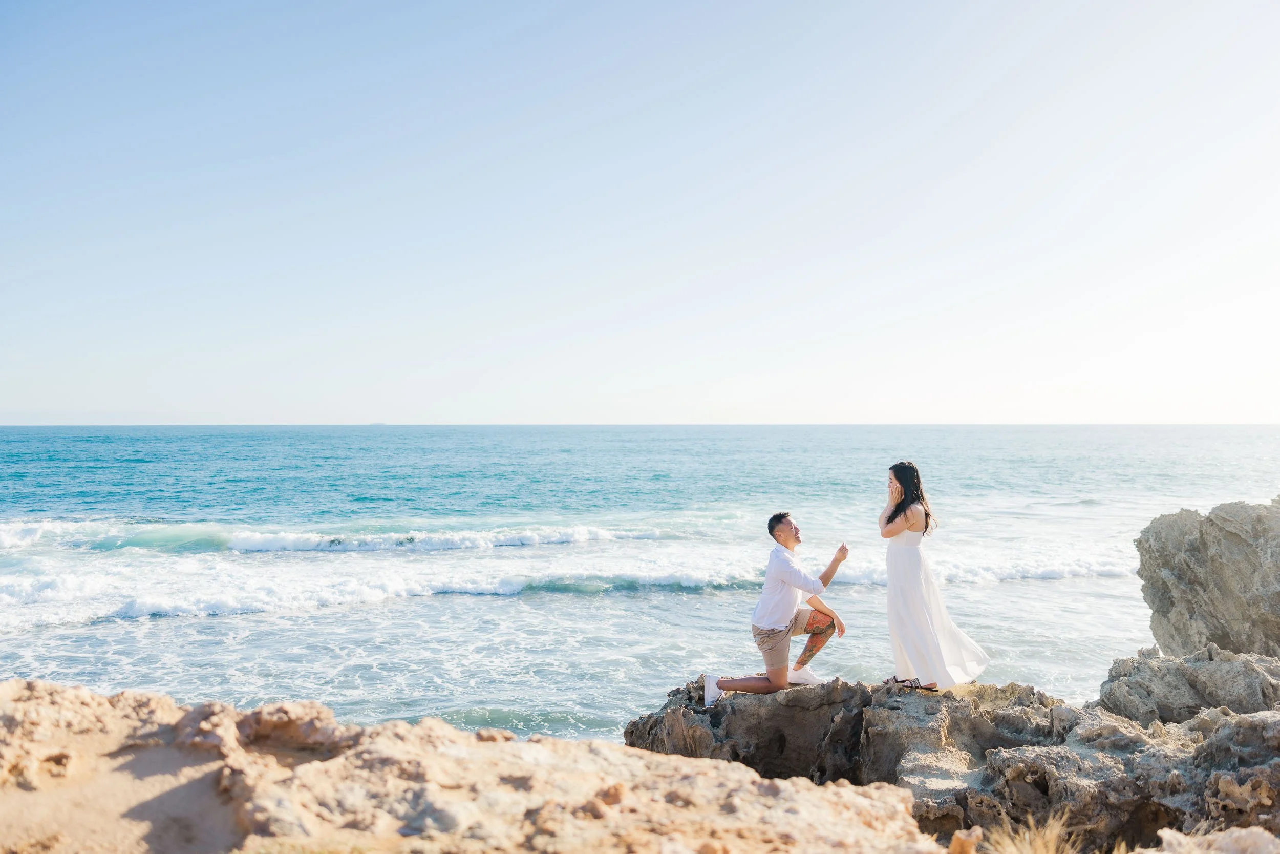 Melbourne Proposal Photography on a rocky beach as a man proposes by the ocean and his partner reacts with surprise.