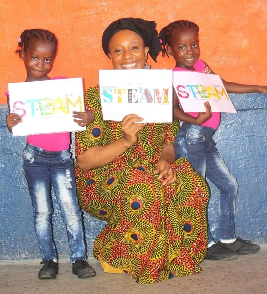 ITAVAC Founder Erica Krutu Davies with two young girls smiling and holding STEAM education signs, representing youth technology and empowerment programs in Brooklyn and Liberia