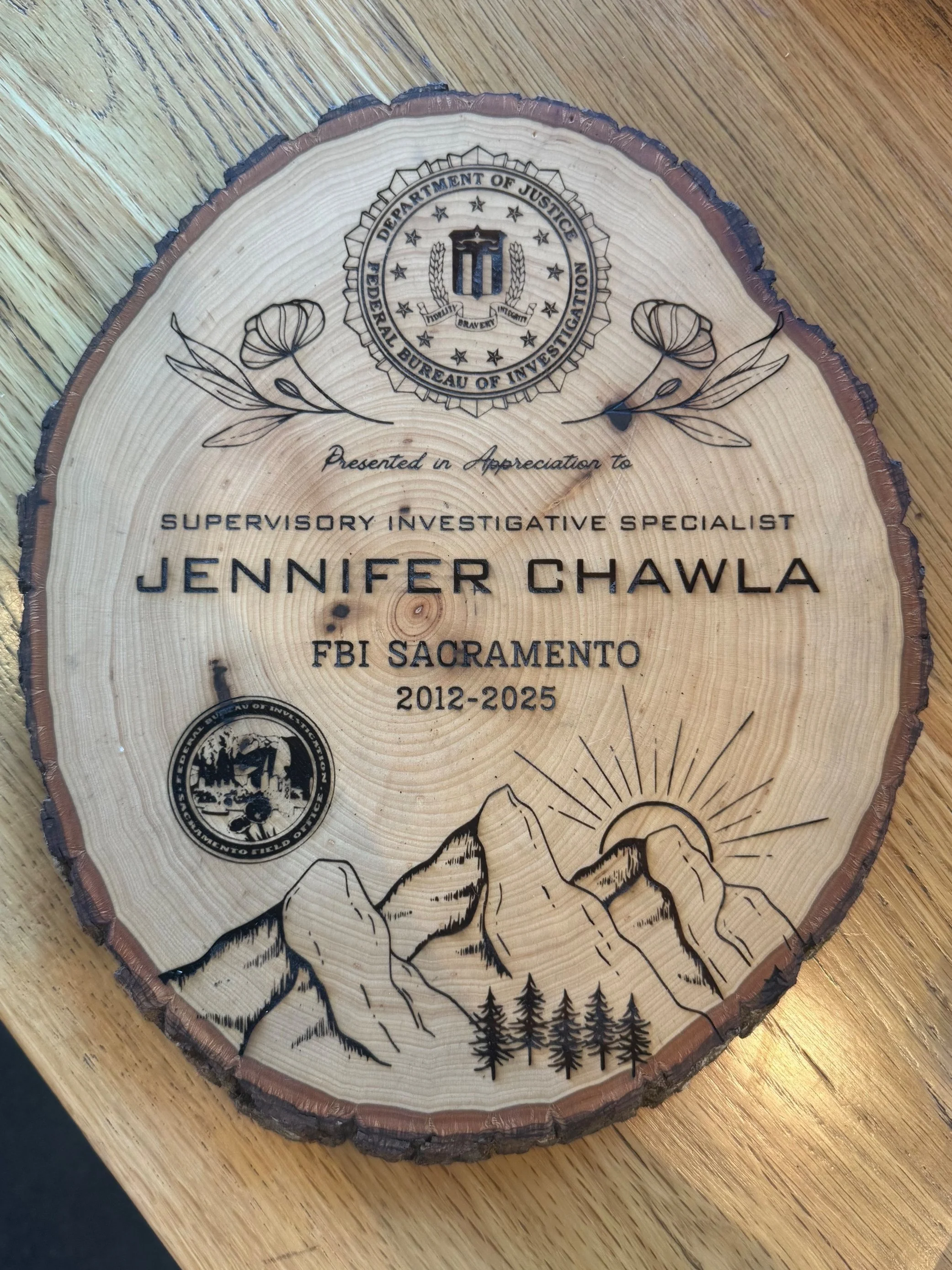 Wooden plaque featuring the FBI seal at the top and a mountain landscape with trees and a sunrise at the bottom, engraved with the name Jennifer Chawla, awarded for a supervisory investigative specialist position in Sacramento from 2012 to 2025.