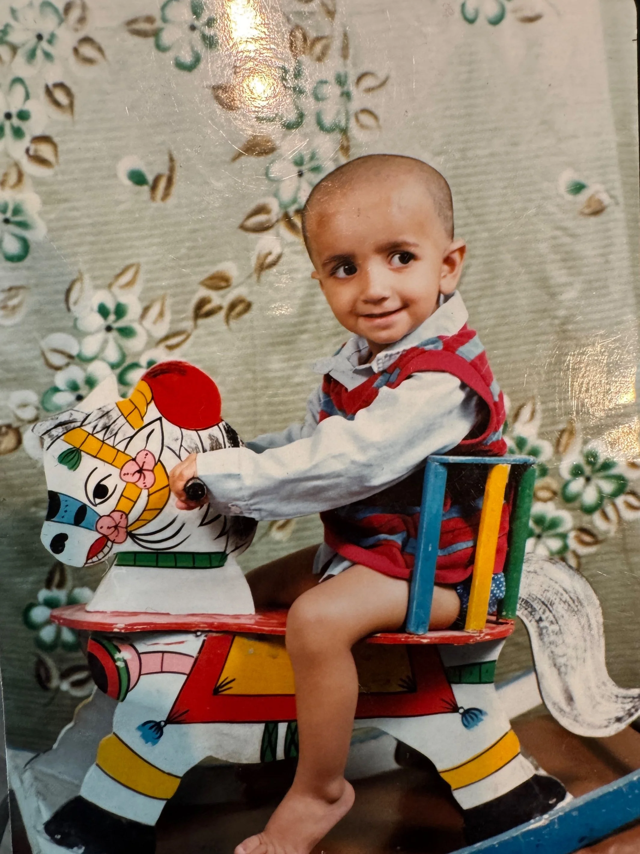 Young child with a shaved head sitting on a colorful wooden rocking horse, holding a paper horse mask, with a floral backdrop.