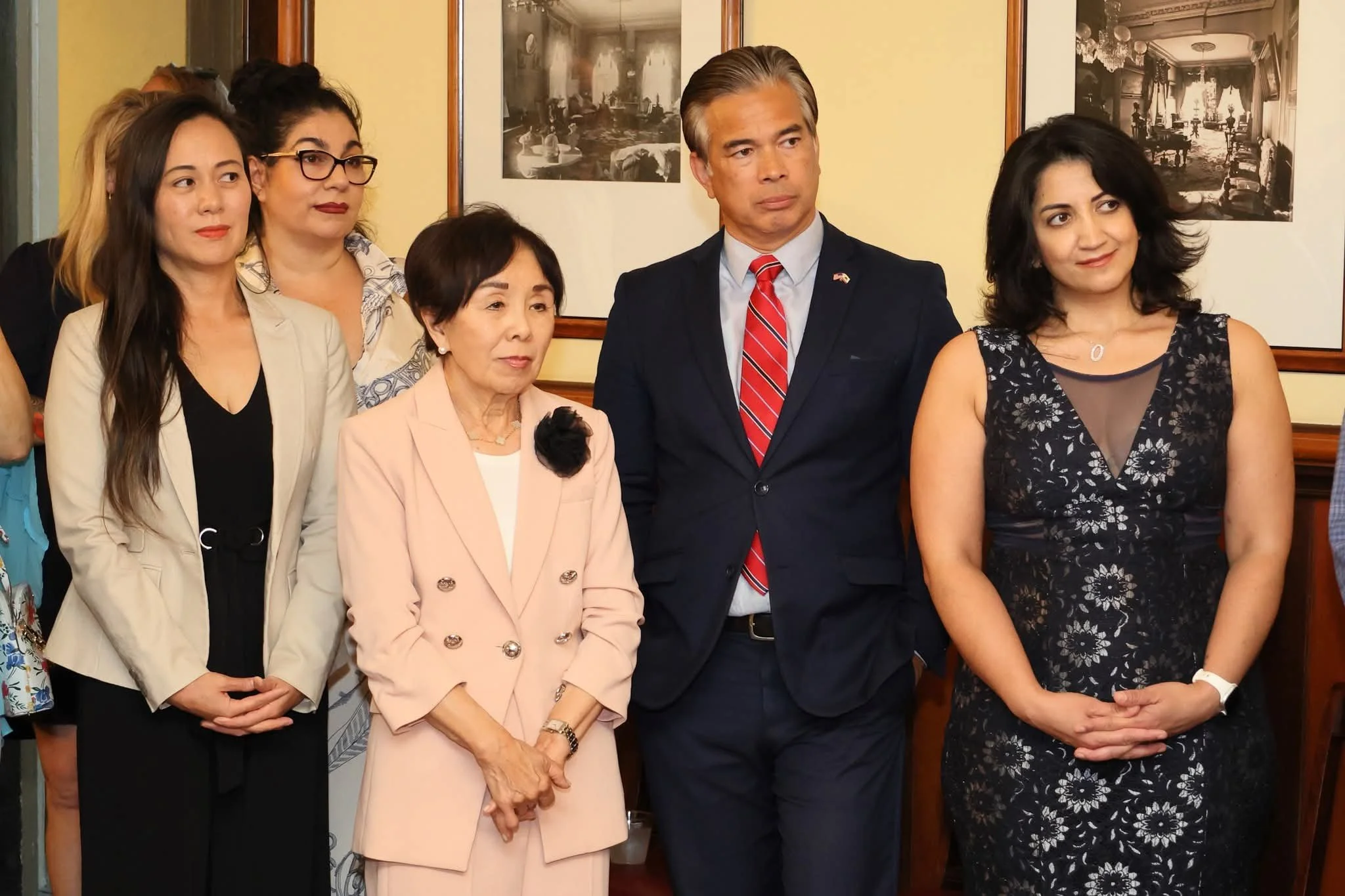 Group of five people standing together indoors, dressed in formal attire, in front of framed black-and-white photographs on the wall.