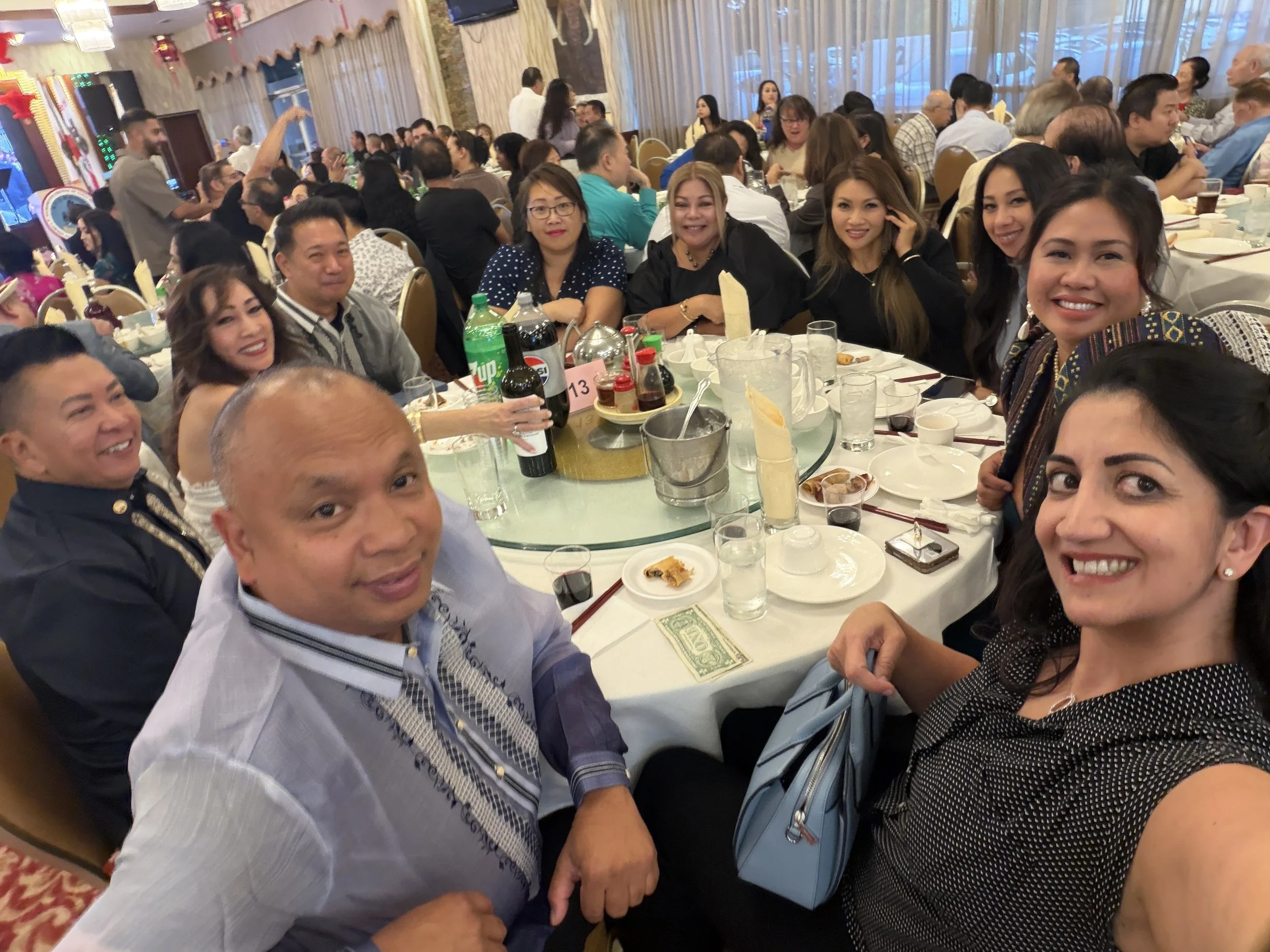 People celebrating at a banquet table with food and drinks, with many more guests in the background, in a decorated banquet hall.