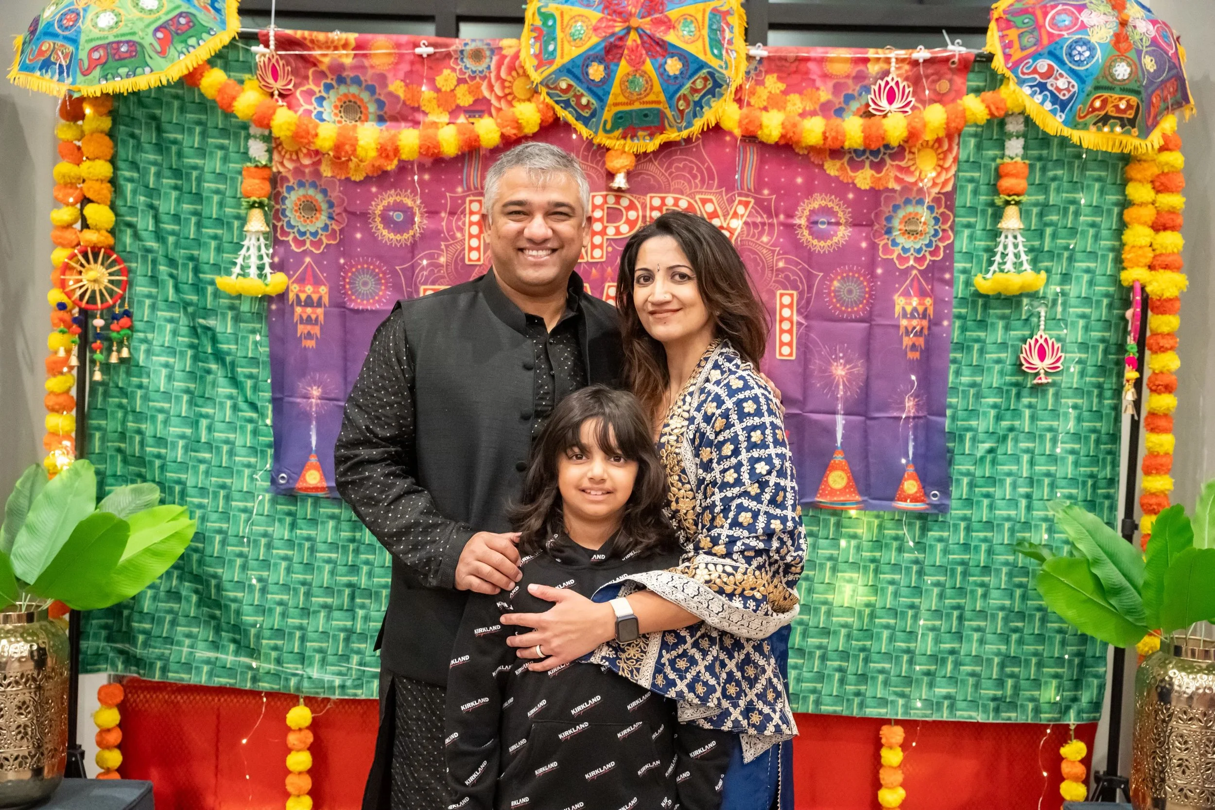 A family of three poses in front of colorful festive decorations including umbrellas and marigold garlands for an outdoor celebration. The man is wearing a black kurta, the woman is in a blue and gold traditional dress, and their daughter is in a black outfit.