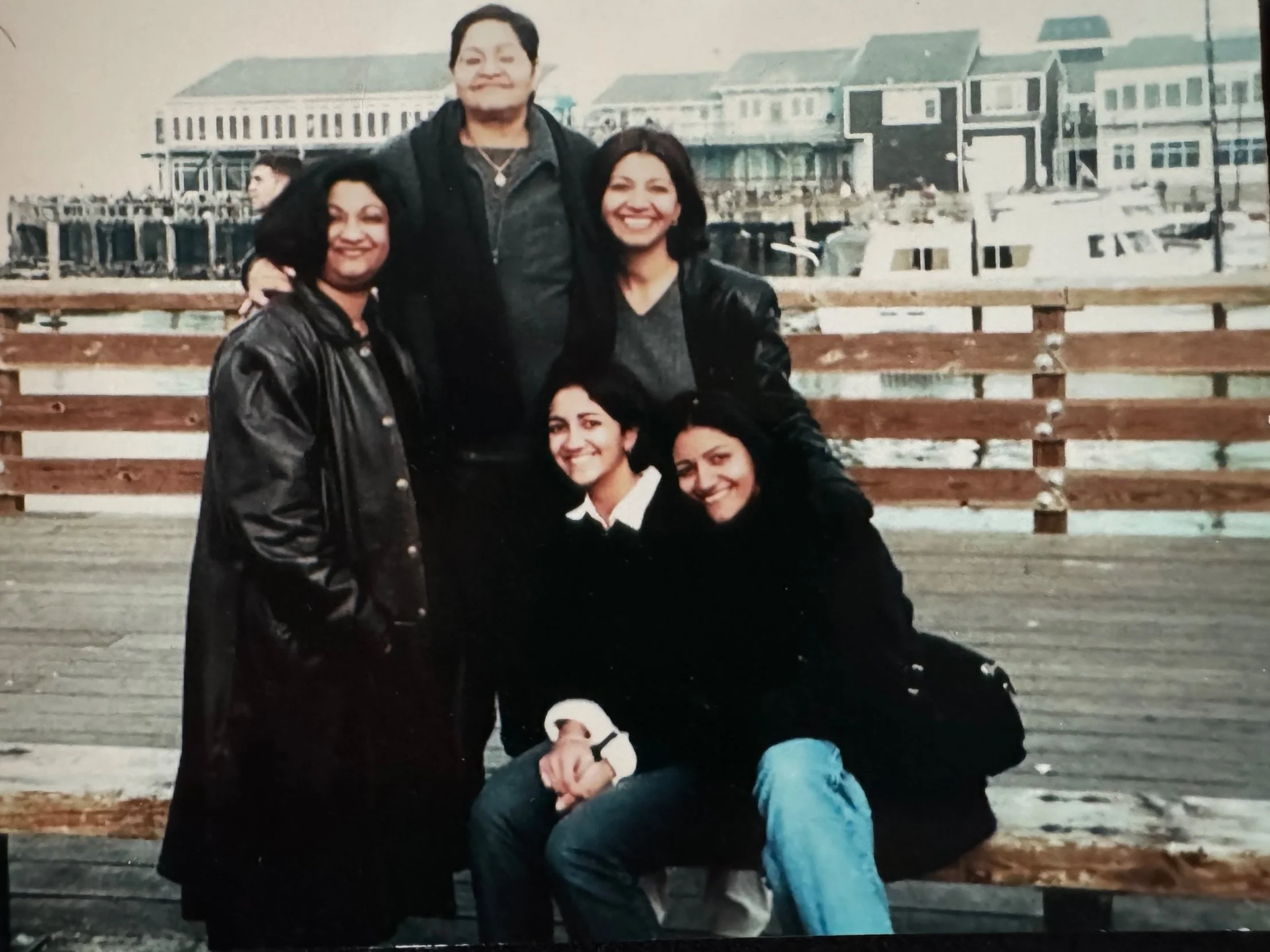 Group of five women posing on a wooden dock with boats and waterfront houses in the background, some women are standing while two are sitting on a bench.
