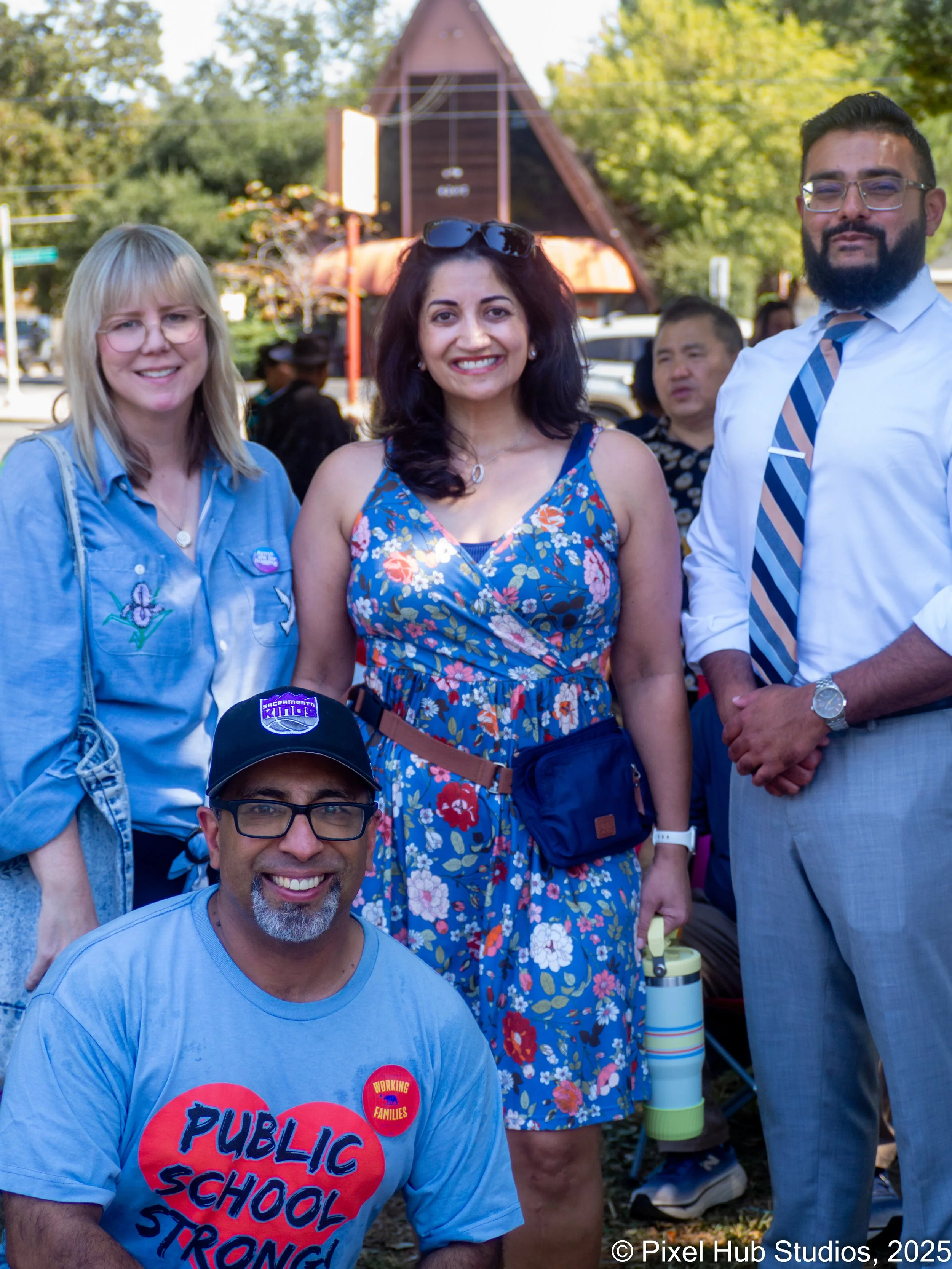 A group of five diverse adults smiling outdoors during a daytime event. One person is wearing a shirt with the text "Public School Strong!" and a button reading "Working Families." Others are dressed casually or professionally, standing in front of a small rustic building and trees.