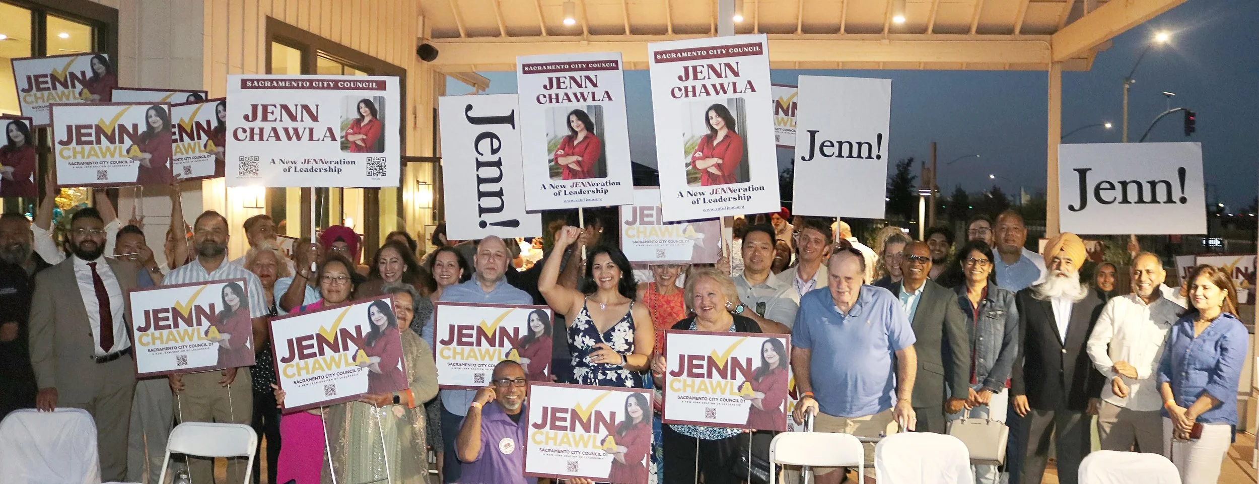 Group of people at a campaign rally holding signs that read 'Jenn Chawla' and 'Jenn!' outside a building during evening.