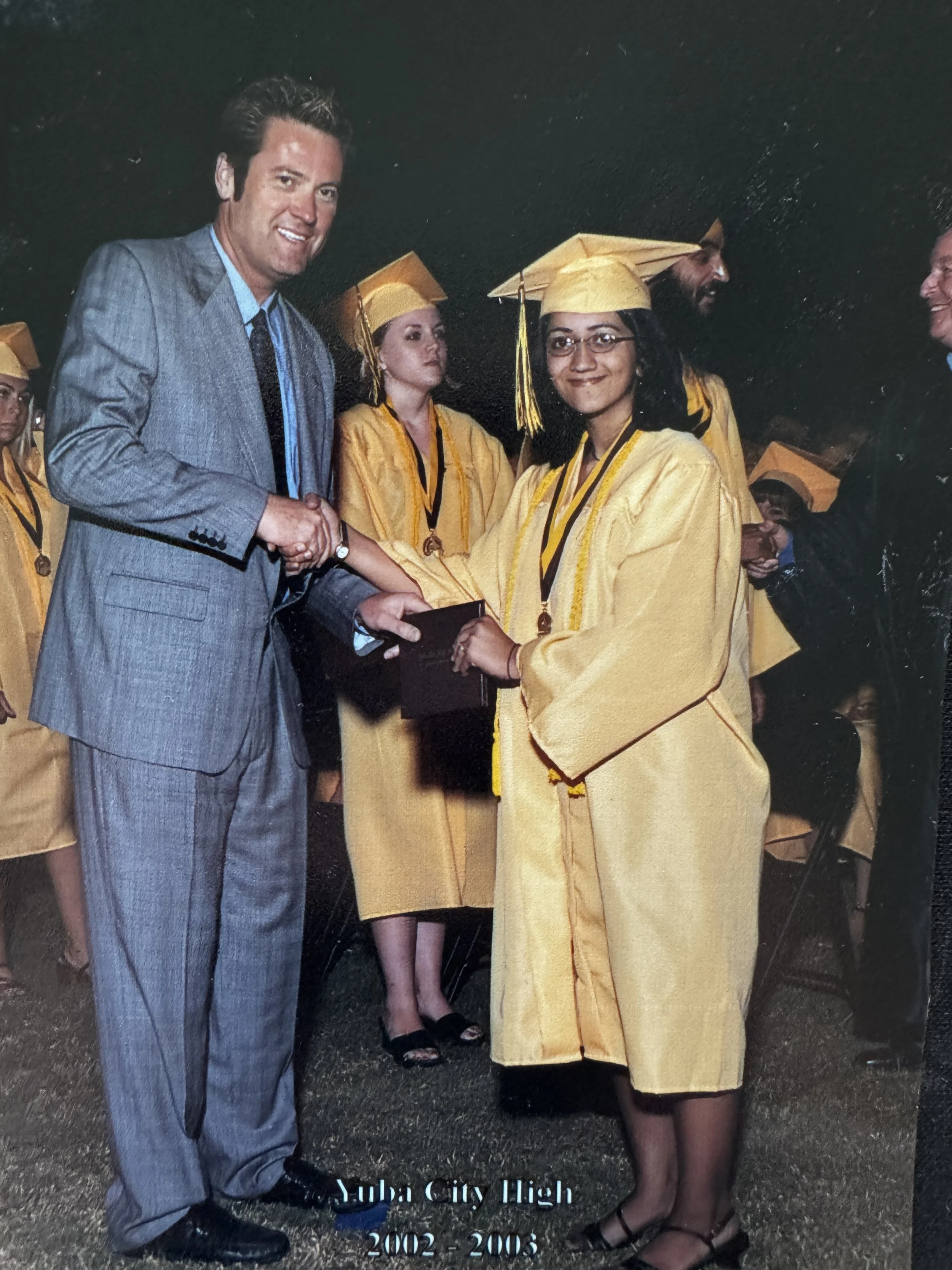 High school graduation ceremony with students in yellow caps and gowns receiving diplomas from a man in a gray suit. Text indicates "Luba City High 2002-2003."
