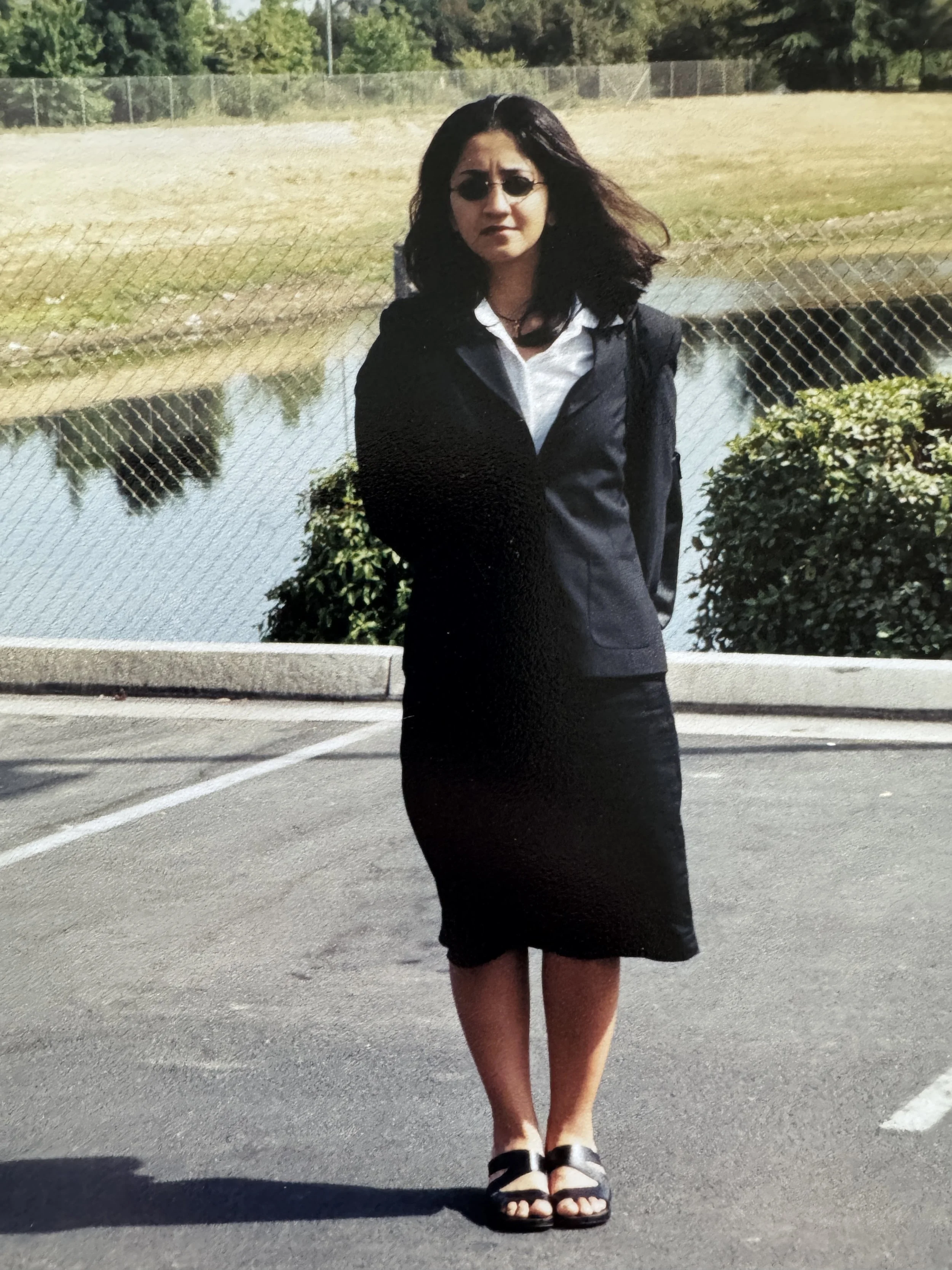 A woman with dark hair wearing sunglasses, a white shirt, black jacket, and black skirt standing in a parking lot near a fence and a body of water with greenery in the background.