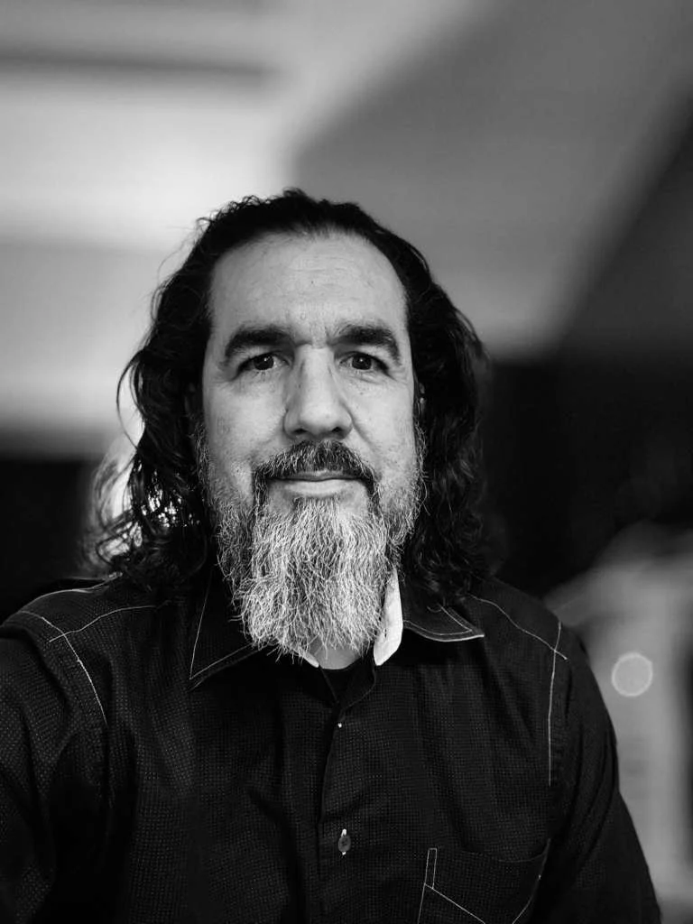 A black and white photograph of Reko Ponton a man with long, dark hair and a full beard, wearing a dark shirt, looking directly at the camera with a serious expression.