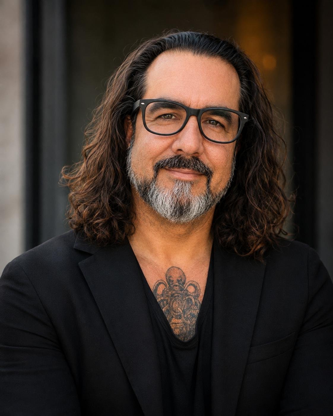 A man with long wavy hair, glasses, and a beard with gray coloring, smiling in front of a dark outdoor background, wearing a black blazer and showing a tattoo on his chest.