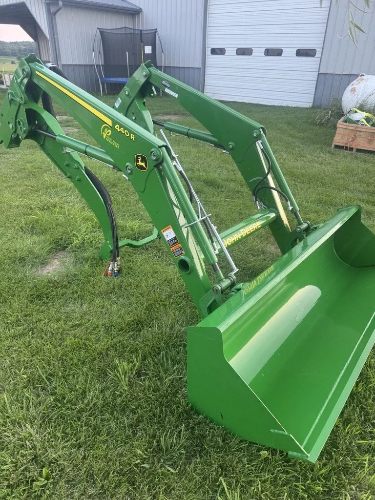 Green John Deere front loader attachment with bucket on grass lawn, with a building and equipment in background.