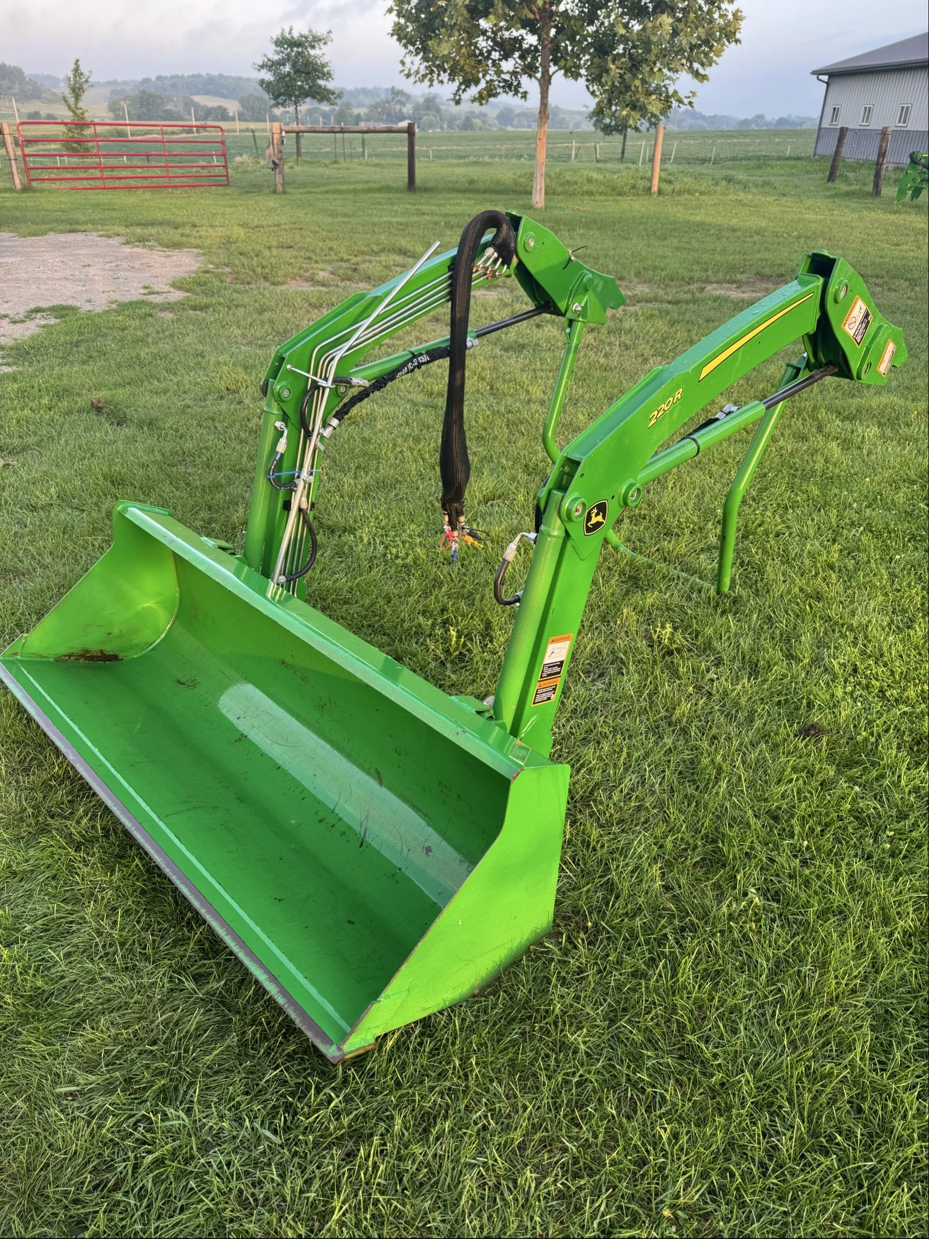 Green John Deere tractor front loader attachment on grass field with trees and farm buildings in background.