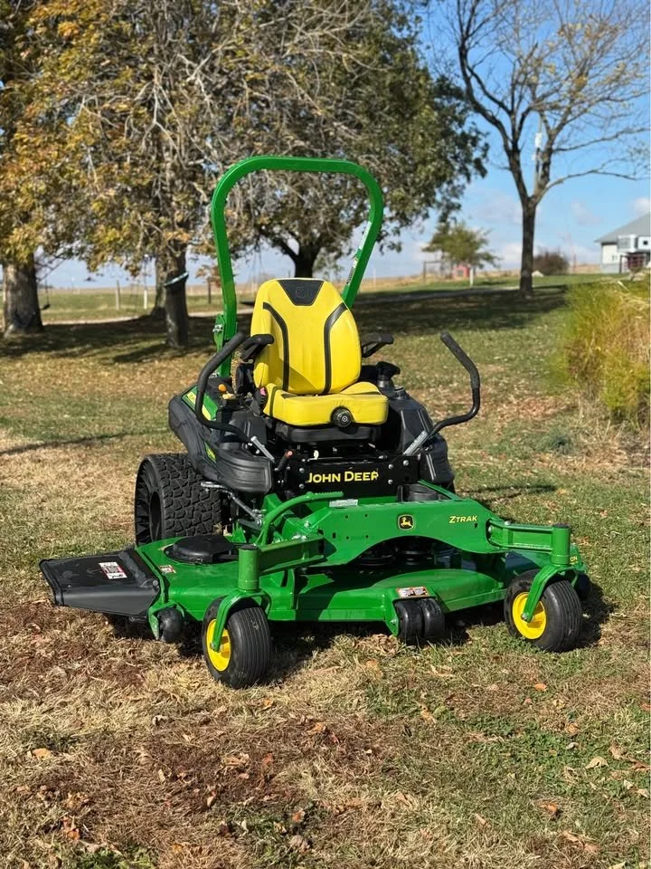 A John Deere lawn mower with a yellow seat and a green roll bar parked on a grassy field with trees and a house in the background.