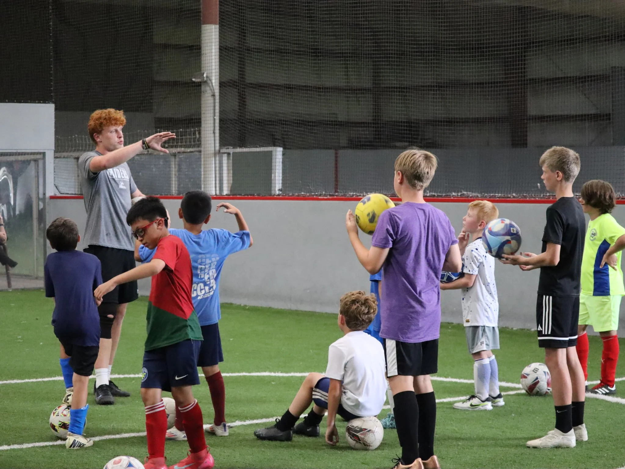 A coach giving instructions to children during a soccer practice indoors. Several children holding soccer balls, some sitting, and others standing on artificial turf.