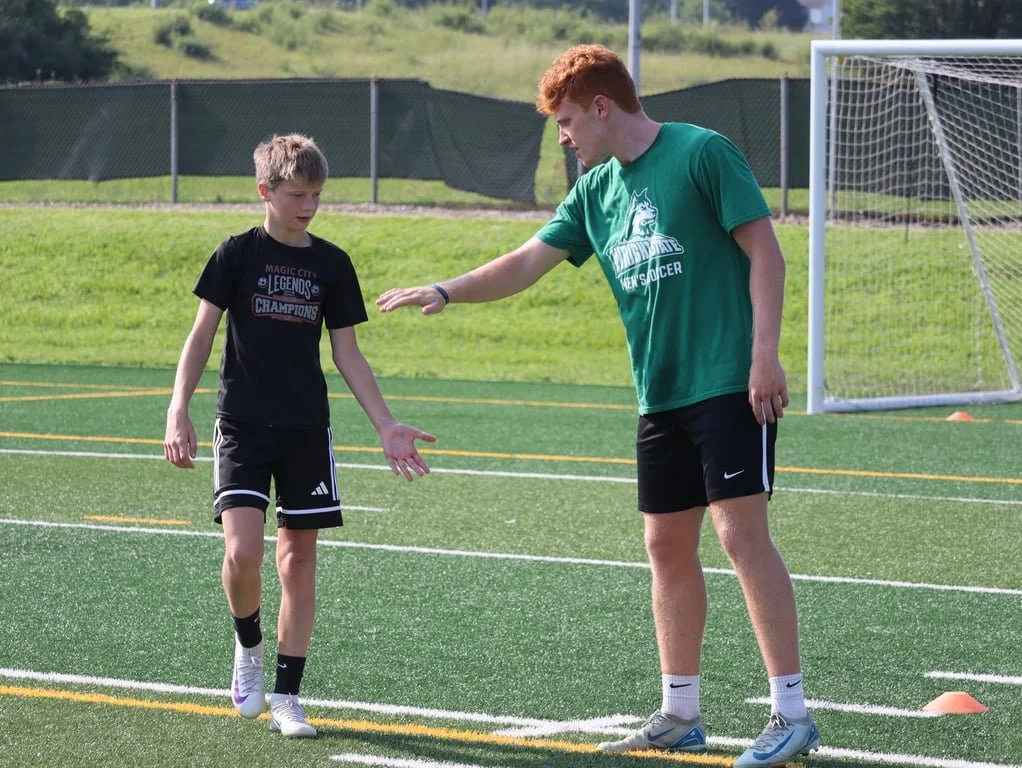 A young boy and a teenage coach practicing soccer on a field with a goalpost in the background. The coach is instructing the boy during a training session.