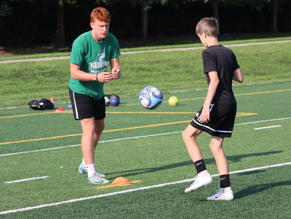 Two boys practicing soccer on a field, one in a green shirt and black shorts, the other in a black shirt and black shorts, with a soccer ball in the air between them.