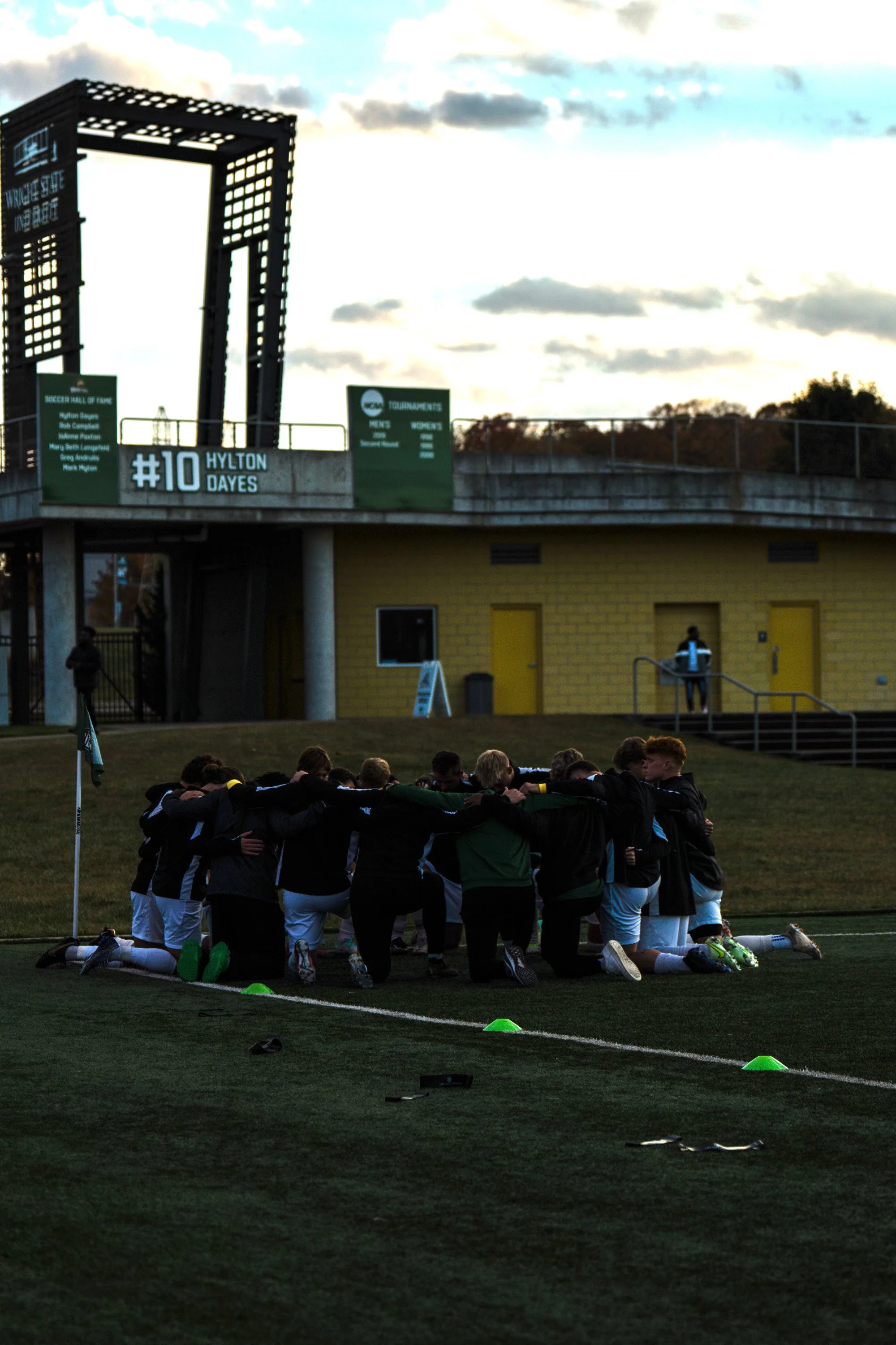 A soccer team kneeling on the field in a huddle during sunset, with players' arms around each other's shoulders, near a corner flag.
