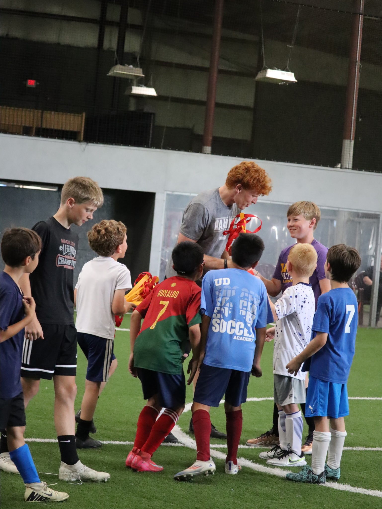 A young man with red curly hair is handing out medals to a group of children on an indoor soccer field. The children are dressed in various sports jerseys and casual clothing, and they are gathered around him, smiling and looking at the medals.