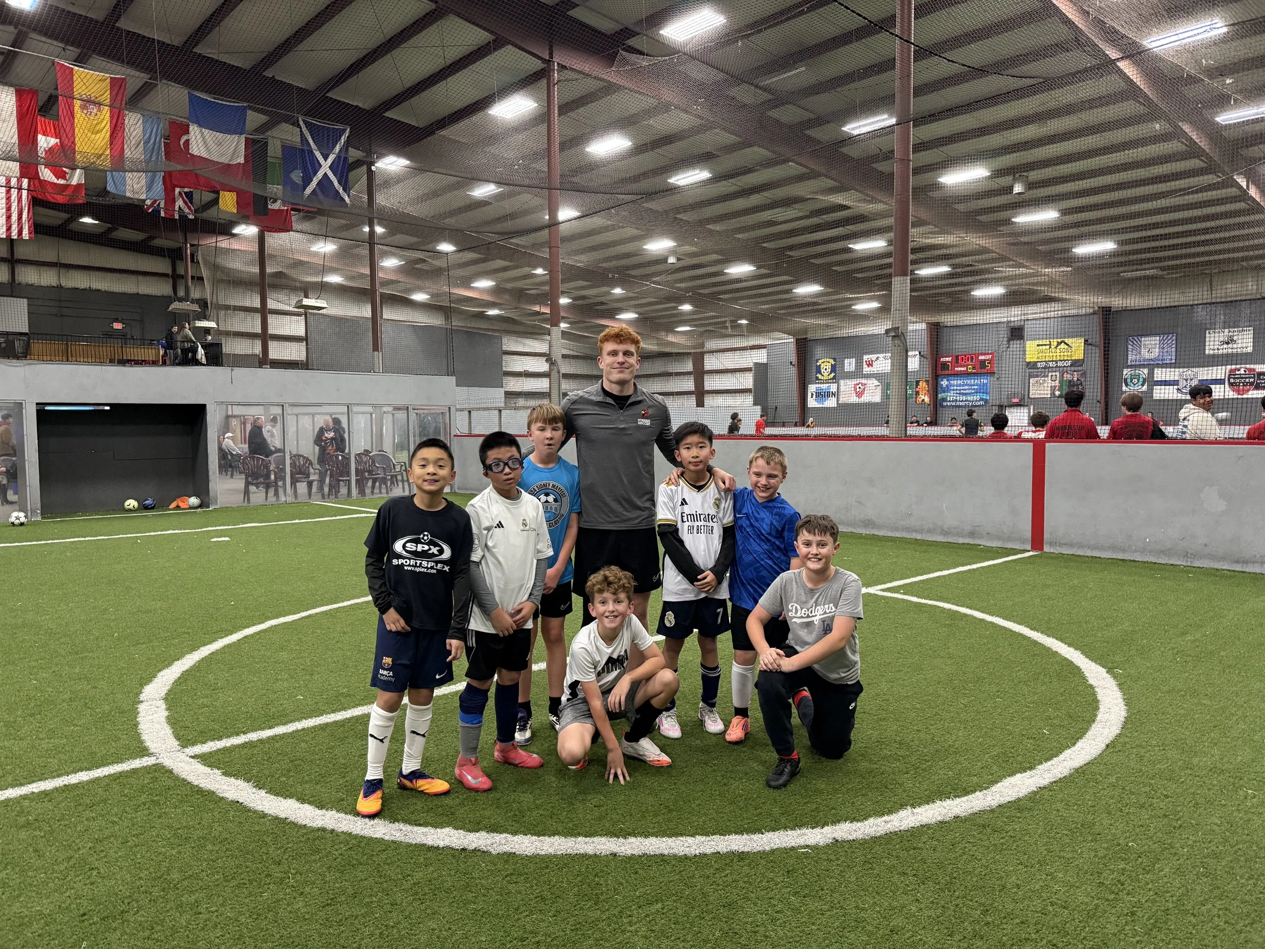 A group of young boys and a coach posing on an indoor soccer field, with spectators and banners in the background.