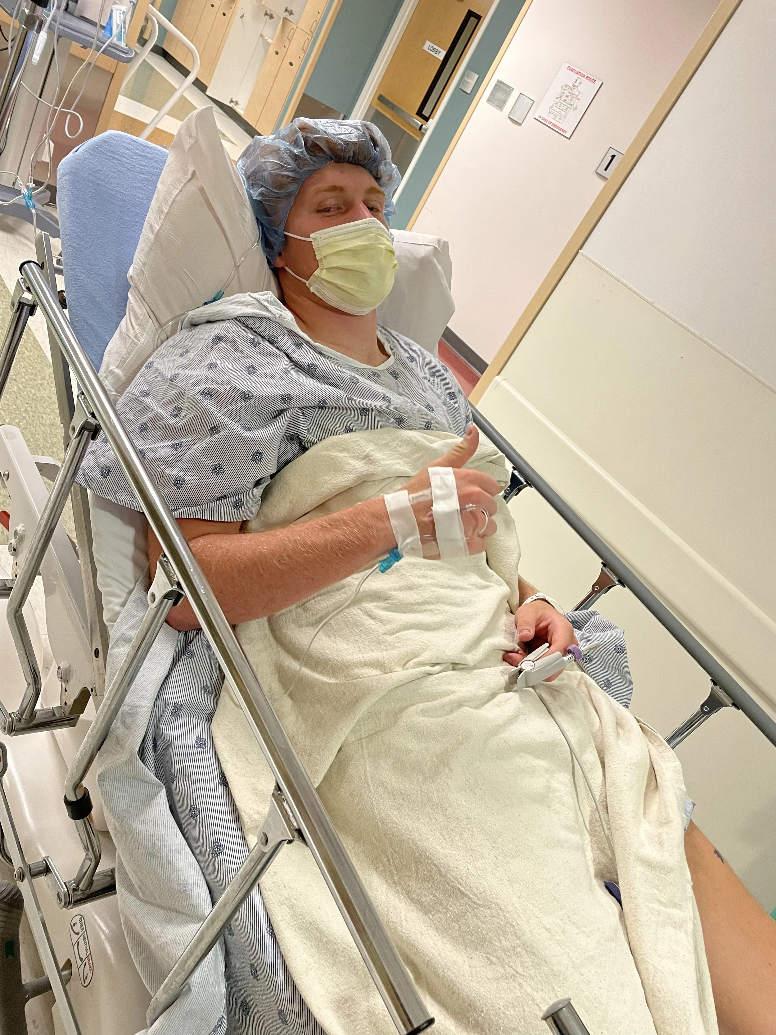 A man lying in a hospital bed wearing a yellow face mask, hair cover, hospital gown, giving a thumbs-up, with IV line attached.