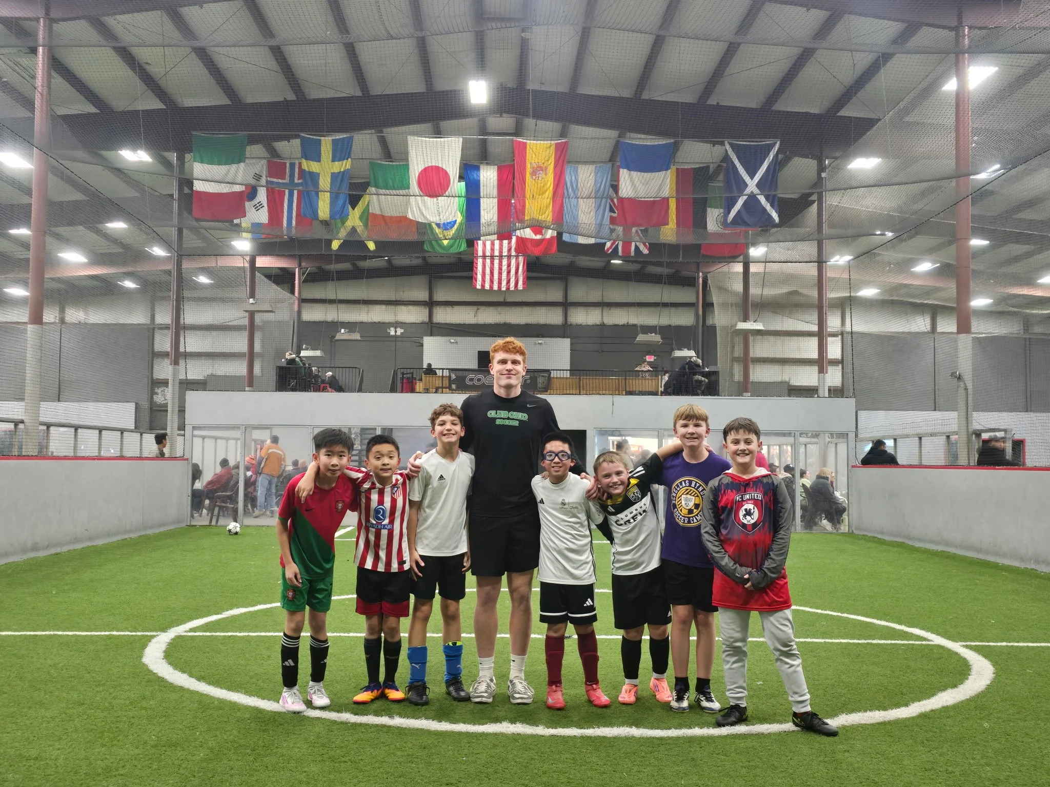 Group of young boys and a coach standing together on an indoor soccer field, smiling, with various national flags hanging overhead, and spectators in the background.