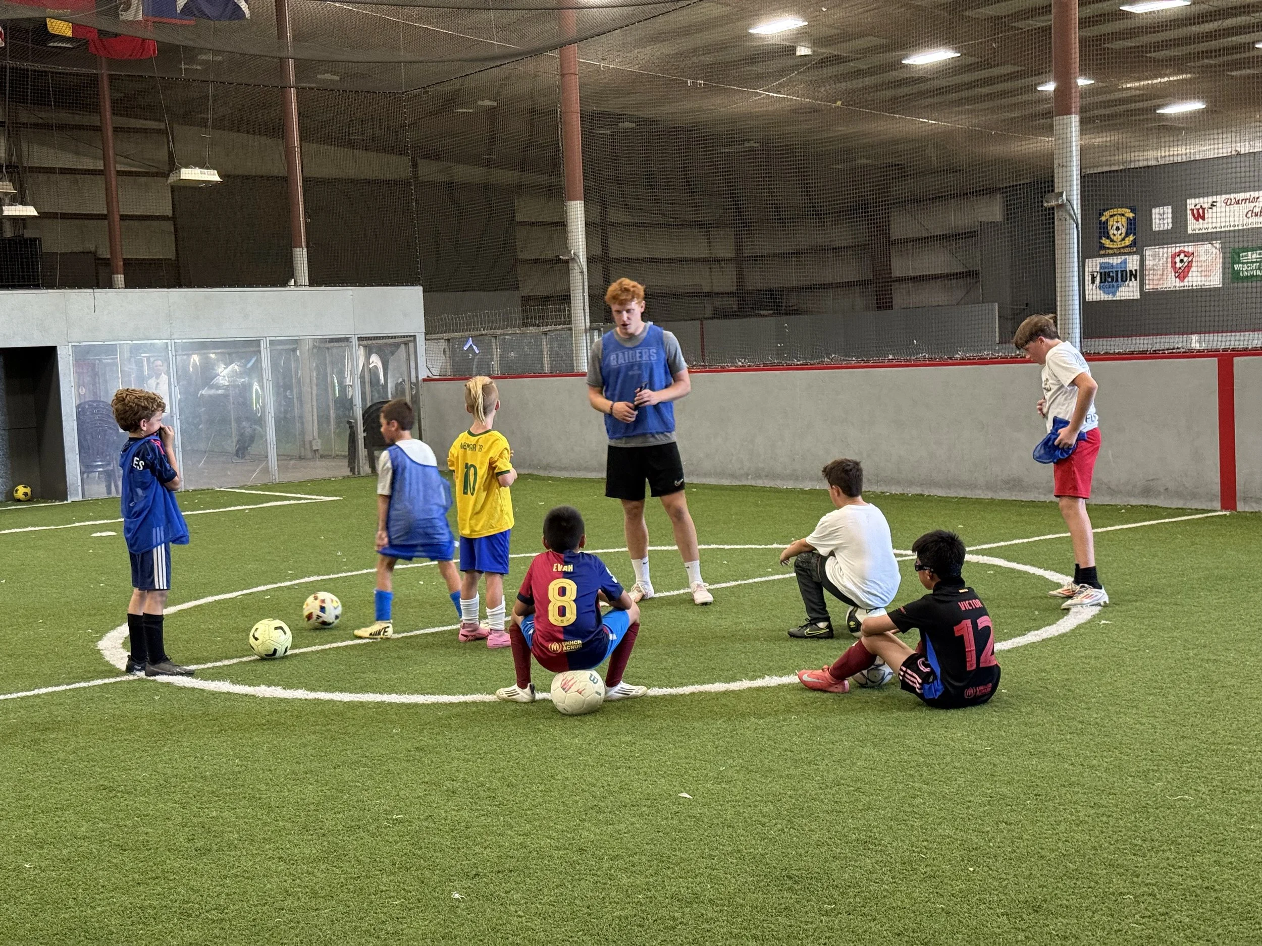 Children and a coach seated in a circle on an indoor soccer field, participating in a soccer practice or discussion, with soccer balls in front of some children.