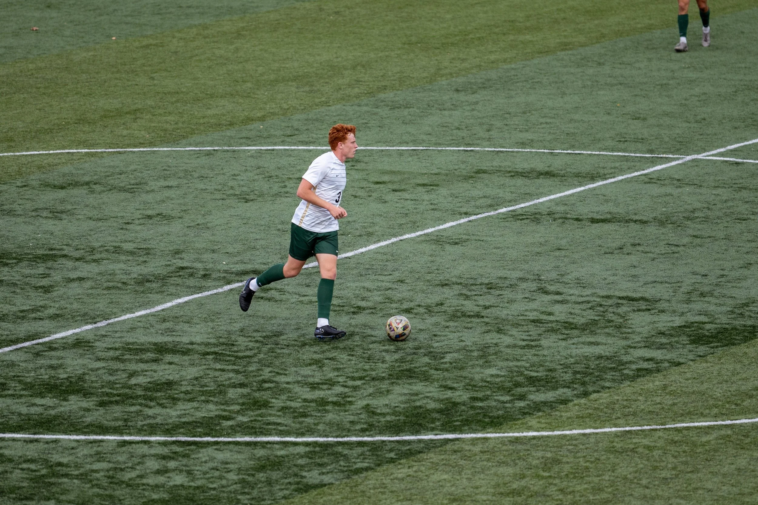 A soccer player with red hair running on a green field with a soccer ball.
