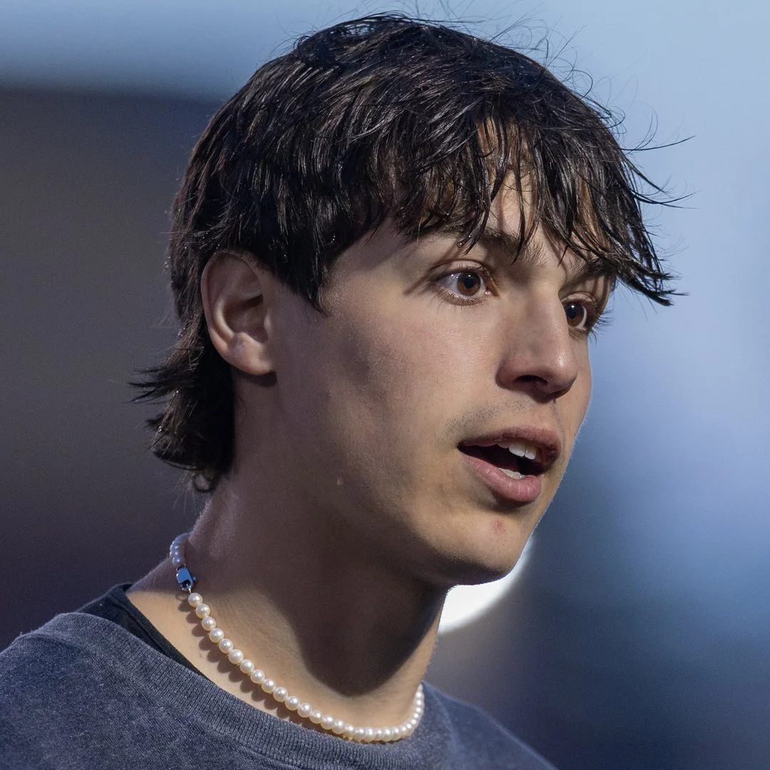 A young man with dark, wet hair, wearing a pearl necklace and a dark shirt, appears to be speaking or reacting to something, with a surprised or intrigued expression.