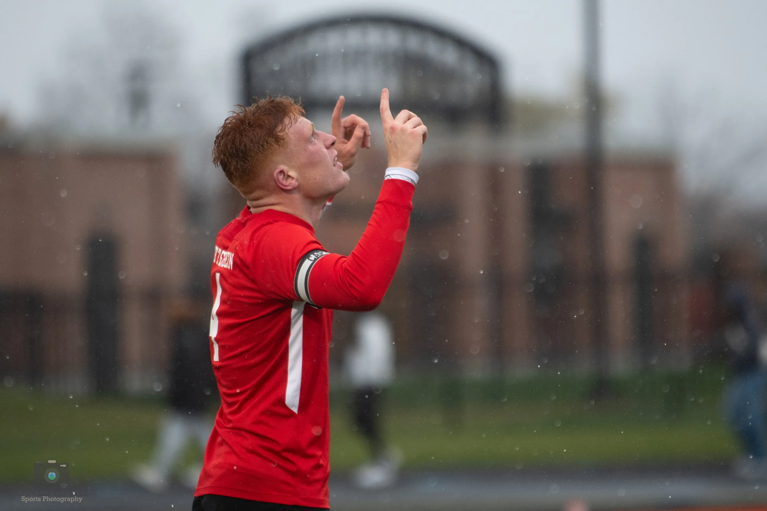 A young man with red hair in a red sports jersey, lifting his hands and looking upward on a rainy day during a soccer match.