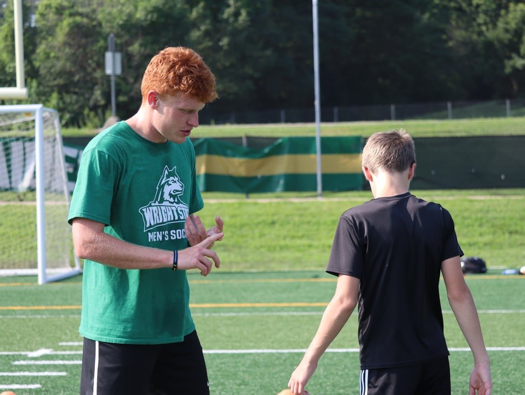 A young man with red hair and a green shirt on a soccer field talking to a boy in a black shirt, with soccer goals and a field in the background.
