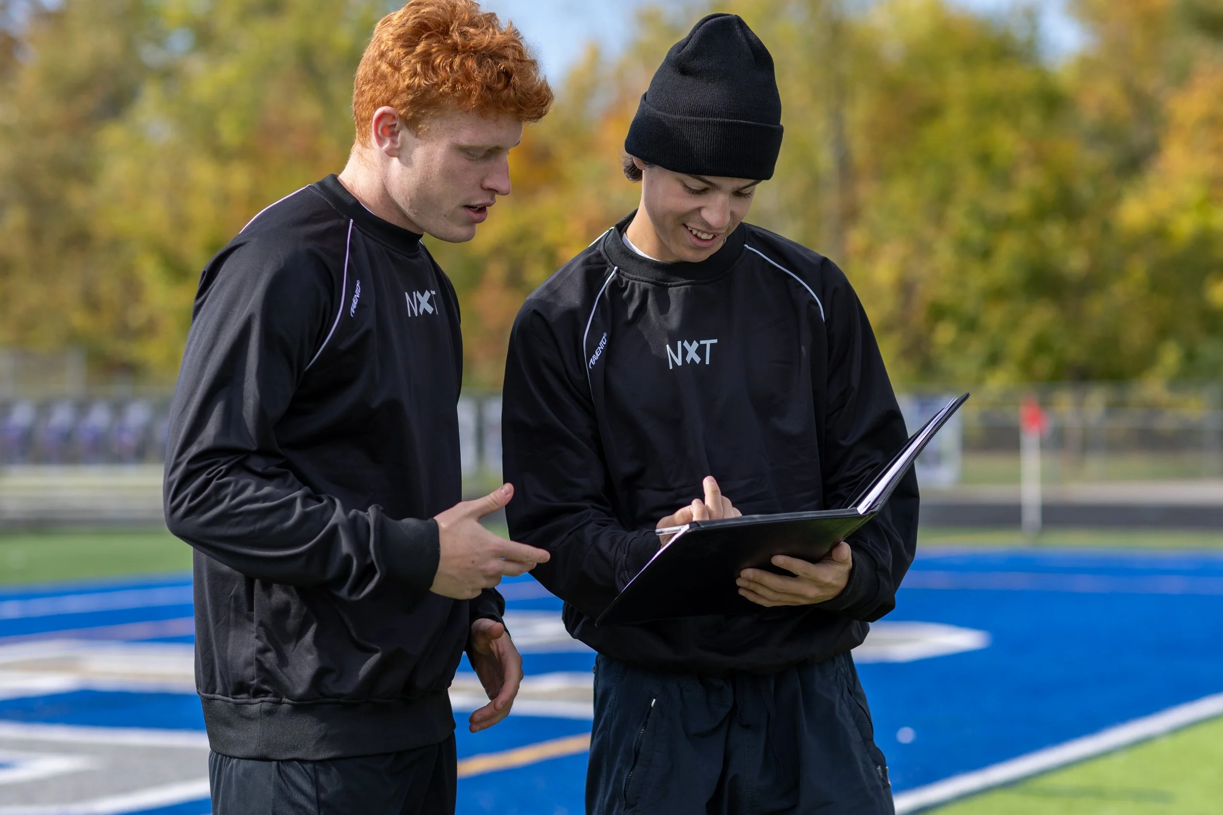 Two young male athletes in black sports jackets with 'NXT' branding standing on a running track, looking at a clipboard together, with a background of trees with autumn foliage.