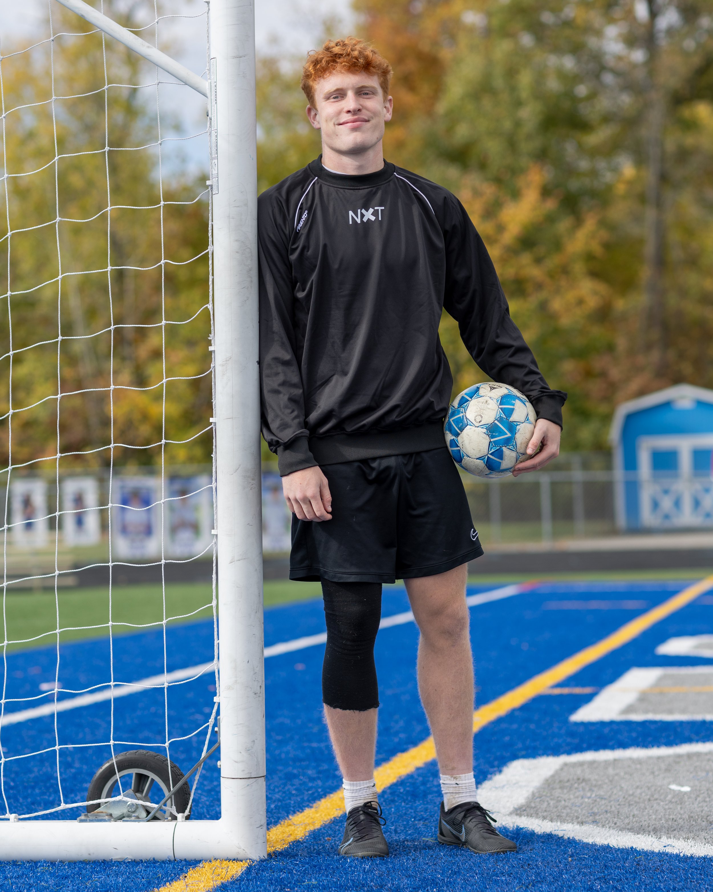 Young man with red hair standing on a blue sports field, leaning against a soccer goalpost, holding a soccer ball, wearing sports attire with a black jacket and shorts, with autumn trees in the background.
