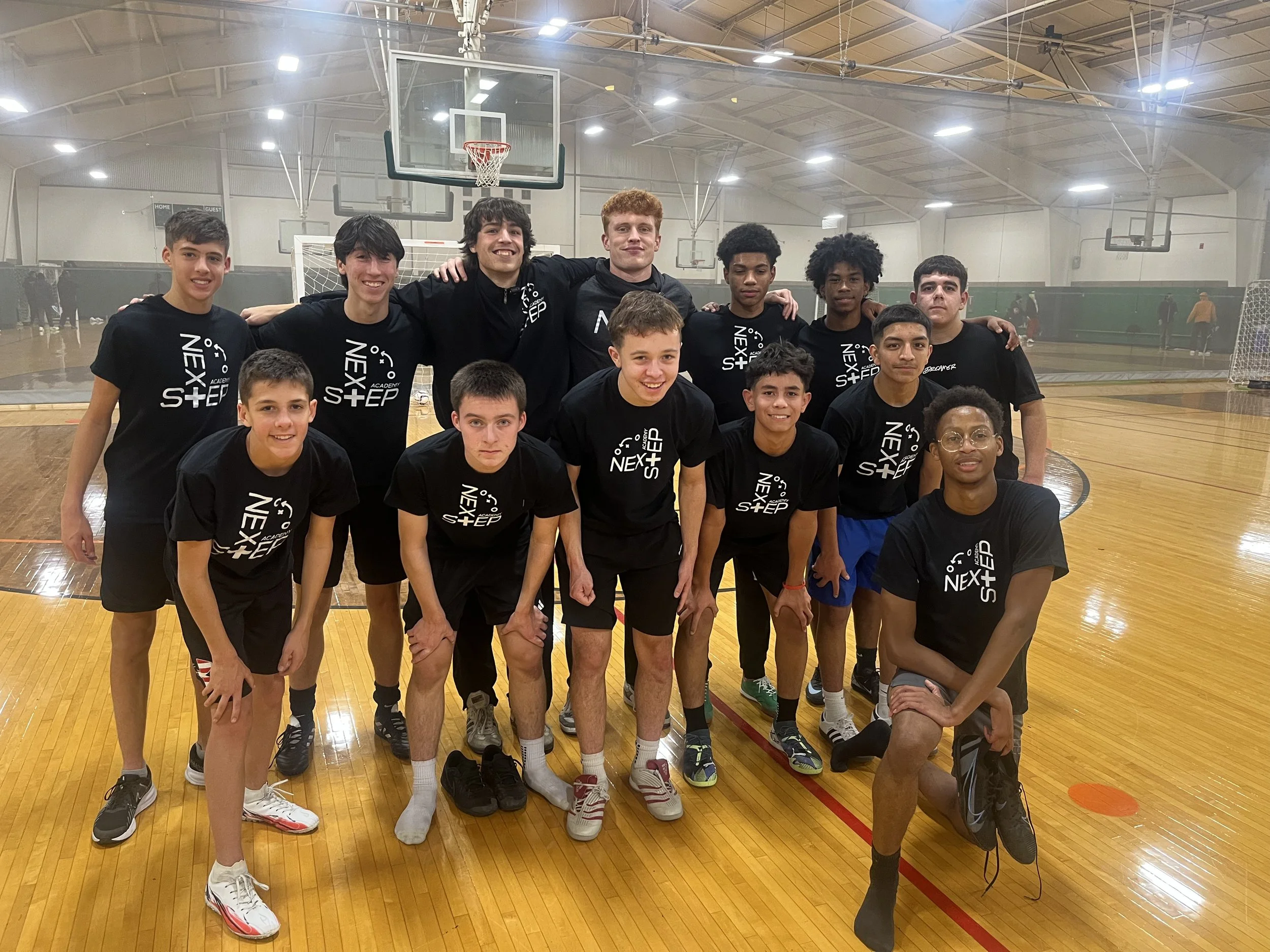 A youth basketball team posing for a group photo on an indoor court. They are wearing matching black T-shirts with the text 'Next Step' printed on them. The team consists of young boys and two older males, likely coaches, standing behind the players.
