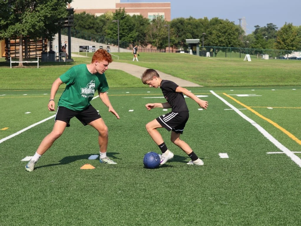 Two boys practicing soccer on a field, with one boy in a green shirt and black shorts facing another boy in a black shirt and black shorts with a blue soccer ball between them.