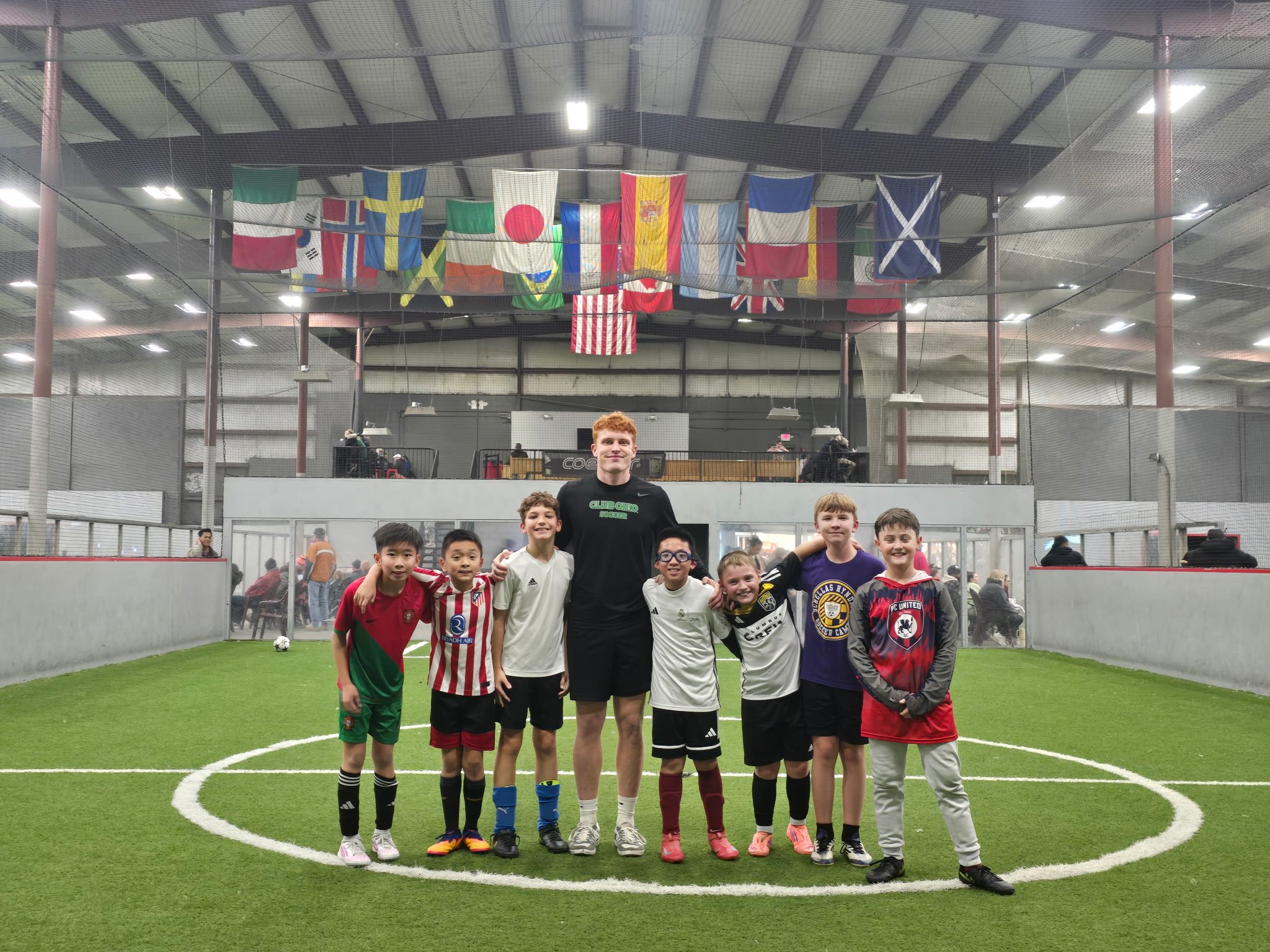 Group of young boys and a coach standing on an indoor soccer field, smiling, with flags hanging above in the background.