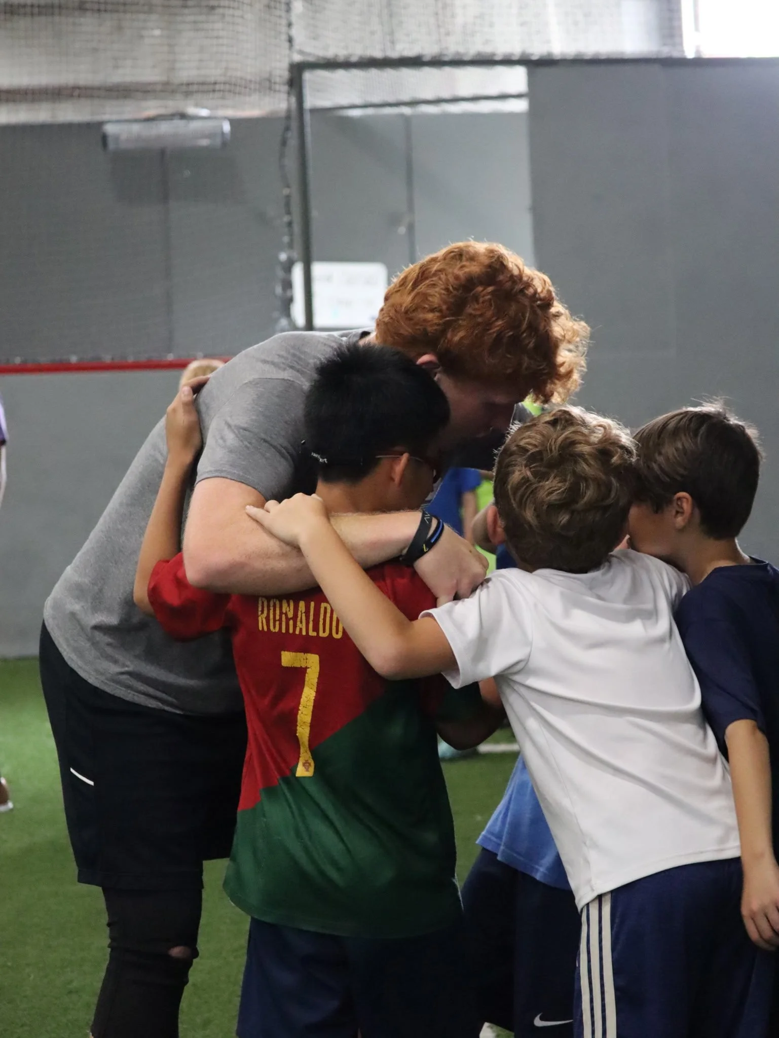 A group of young children and a coach hugging each other in an indoor sports facility.