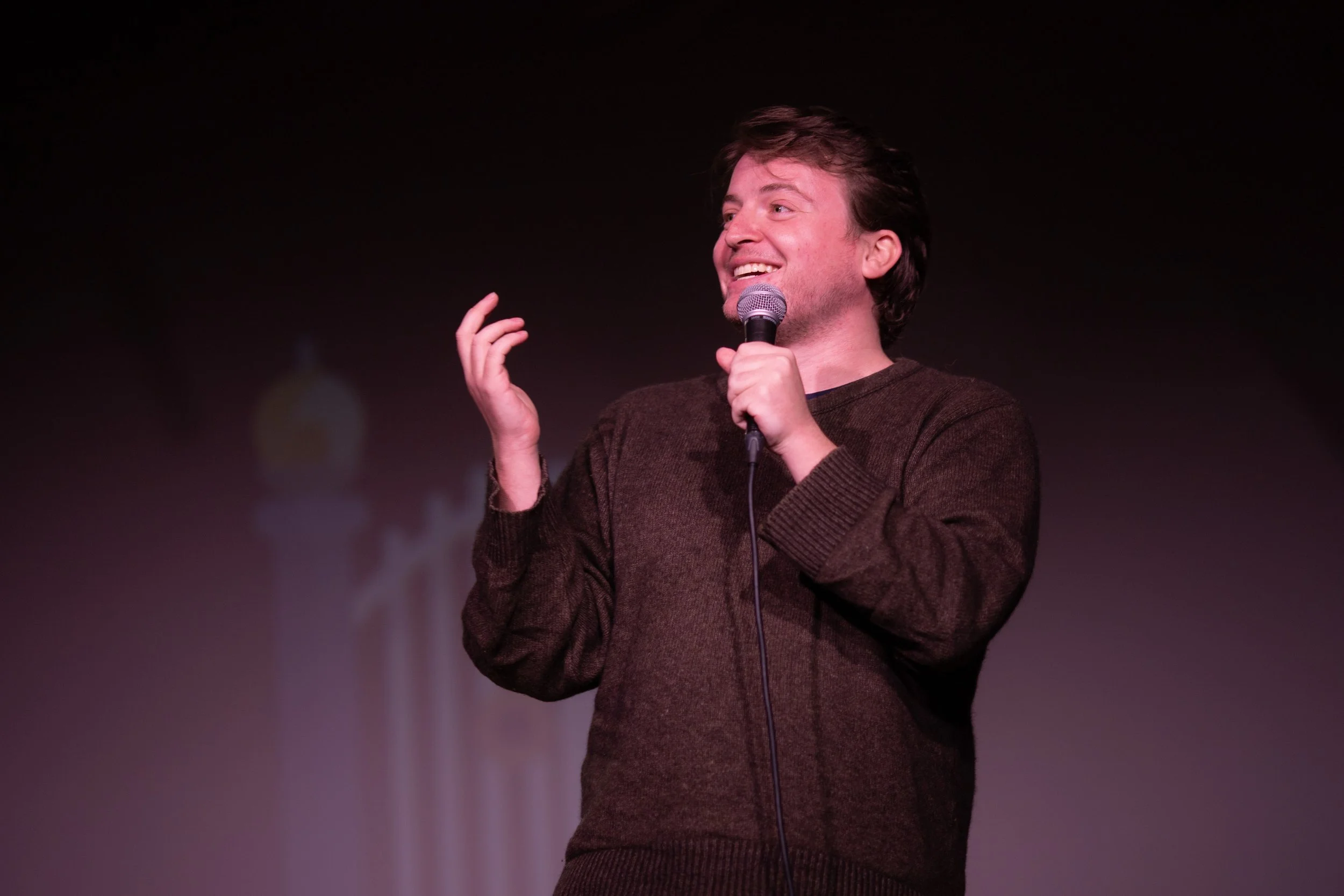 A man on stage holding a microphone, smiling, wearing a brown sweater, with a blurred background.