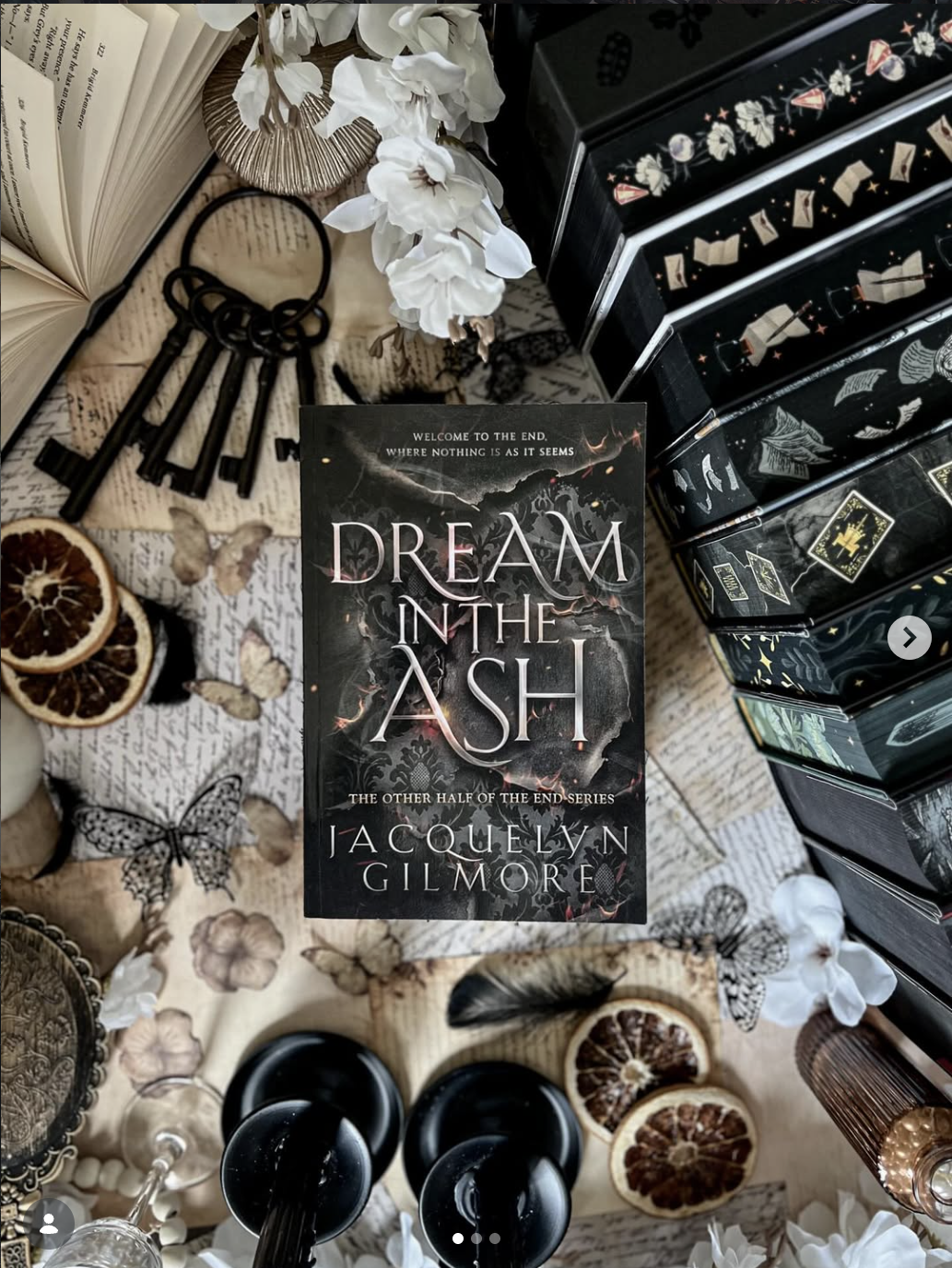 A book titled "Dream in the Ash" by Jacquelyn Gilmore is on a table surrounded by decorative items including dried citrus slices, white flowers, black keys, and various jewelry pieces, with stacked black boxes with floral and book motifs on the side.