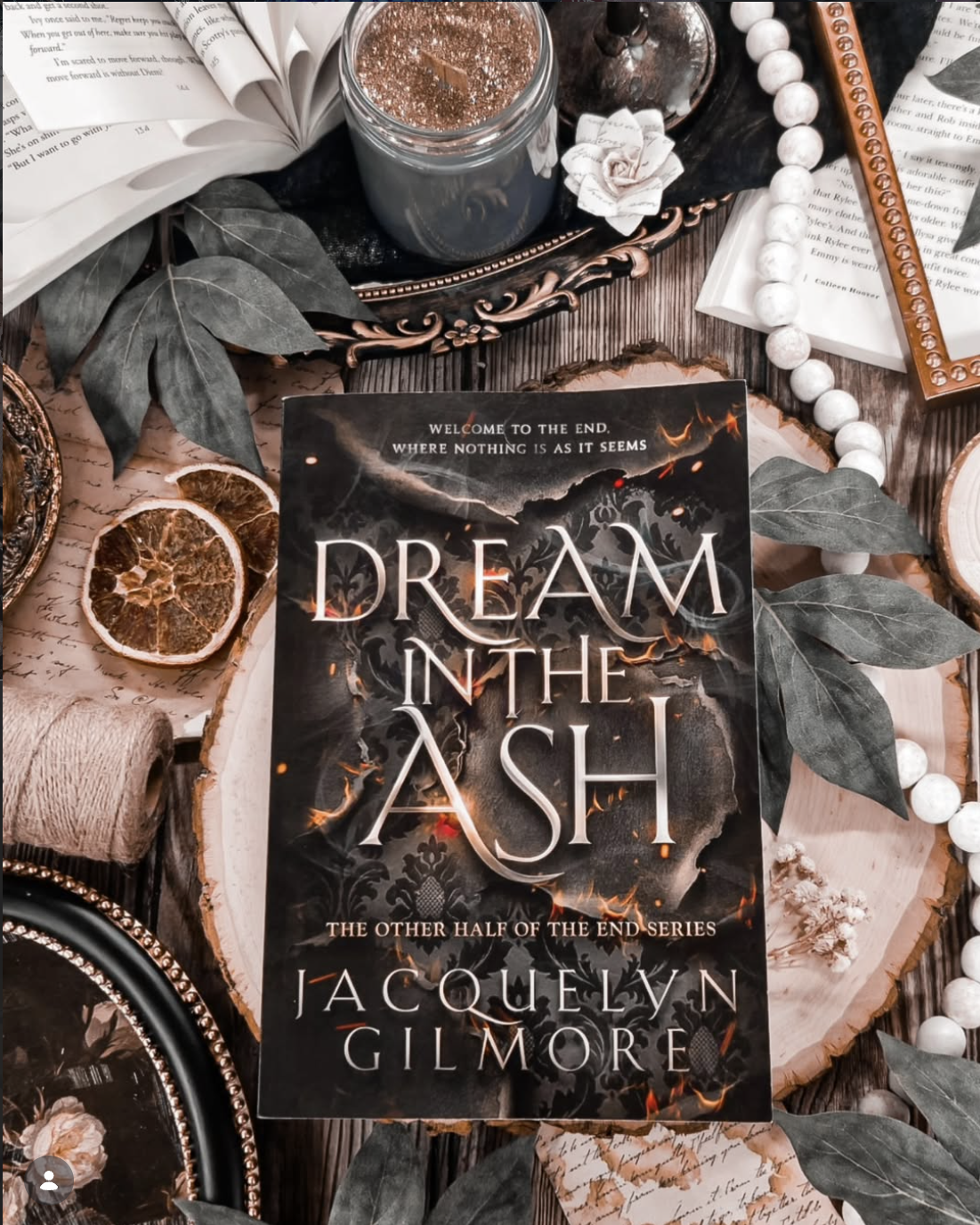 A book titled 'Dream in the Ash' by Jacquelyn Gilmore placed on a decorative tray, surrounded by dried orange slices, green leaves, a jar with a metallic lid, a string of white beads, printed paper, a spool of twine, and other decorative items on a wooden surface.