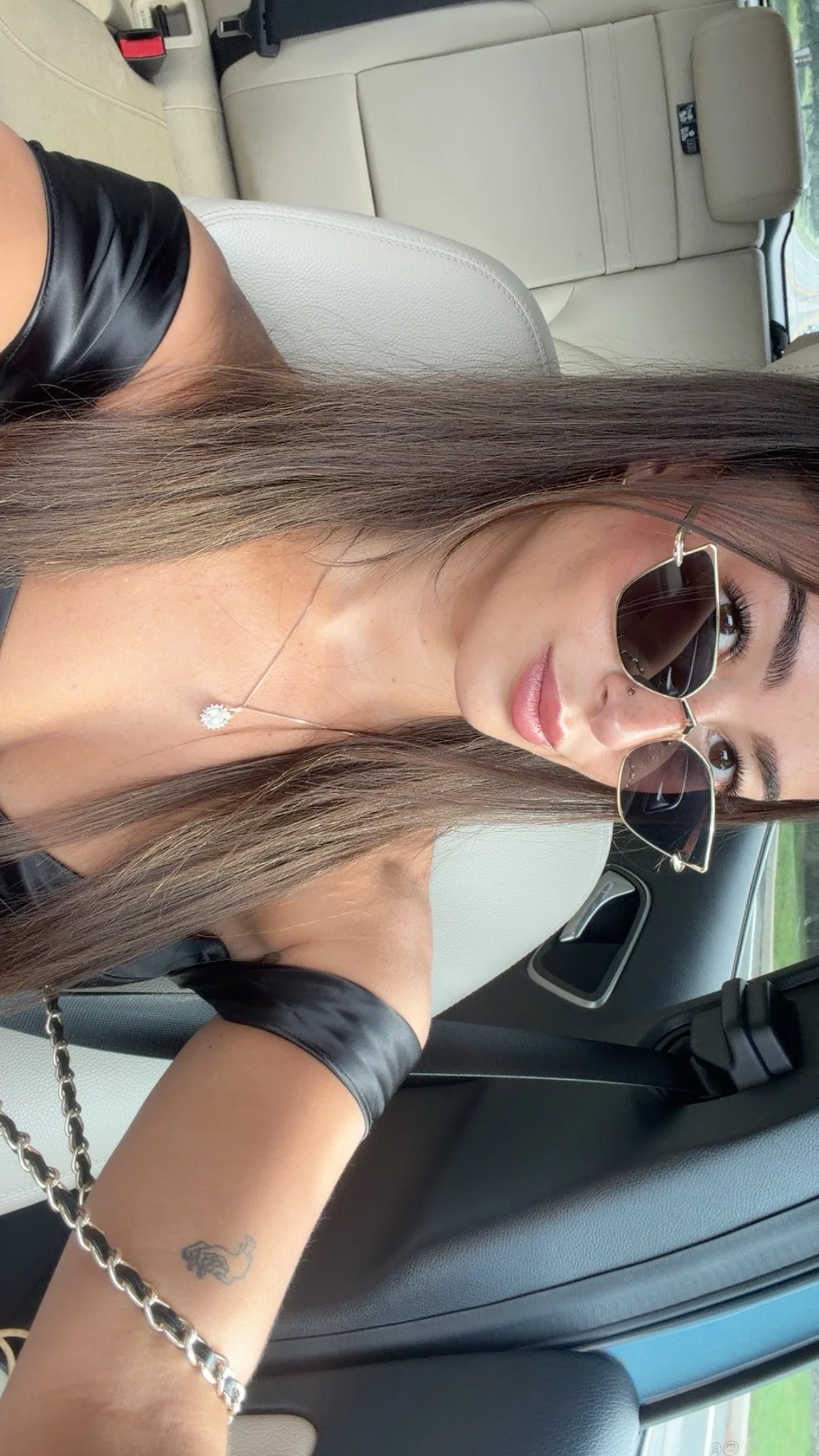 A woman with long brown hair, wearing sunglasses, a silver necklace with a diamonds-shaped pendant, an orange tattoo on her wrist, and a black top, sitting in a car with beige leather interior.