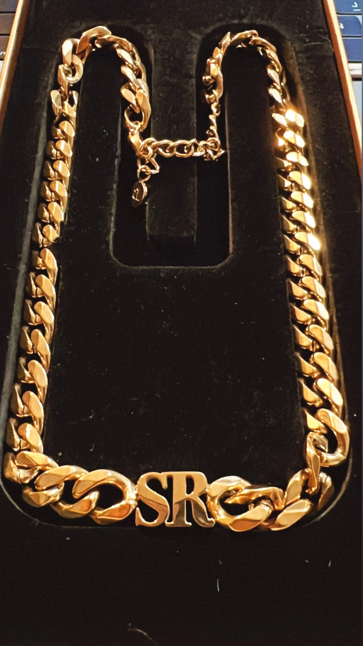 Gold chain necklace with the initials 'SR' at the bottom, inside a box with a black velvet background.