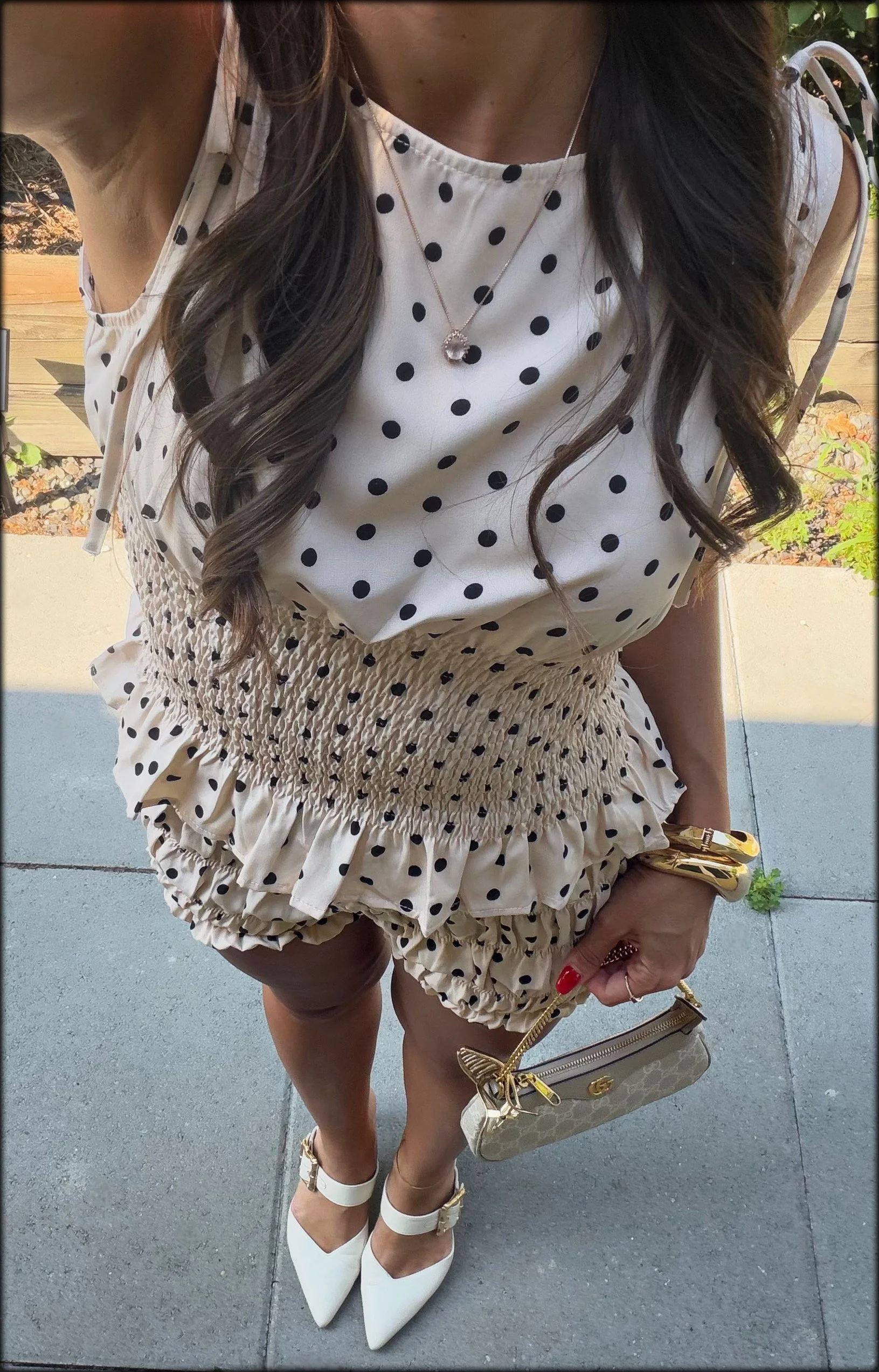 Woman wearing a white polka dot top and shorts, holding a beige handbag, standing outdoors on a paved area.