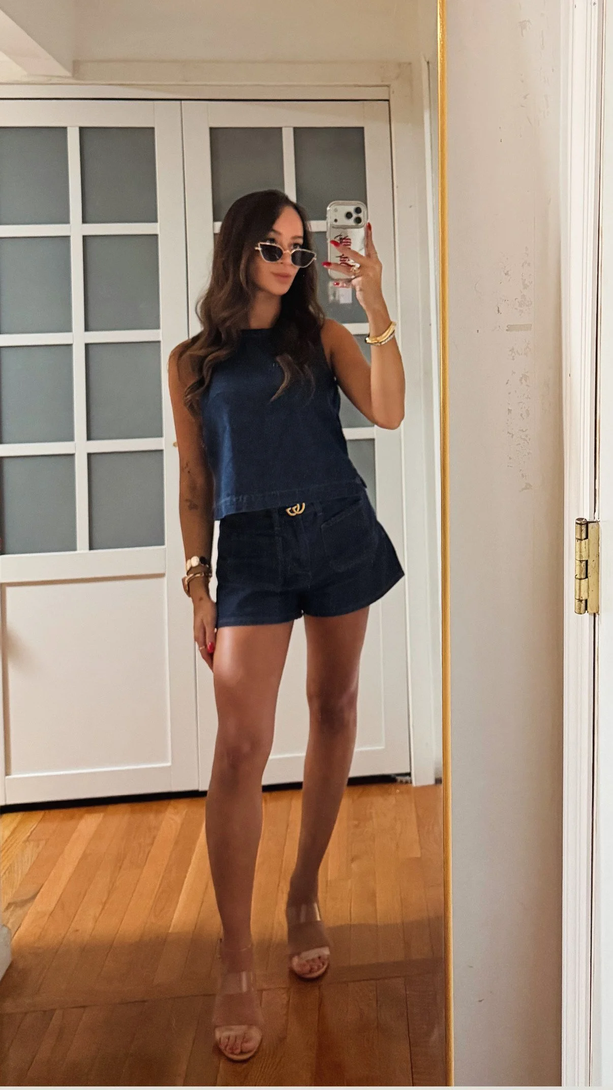 A woman taking a mirror selfie in a room, wearing sunglasses, a sleeveless top, denim shorts, and sandals.