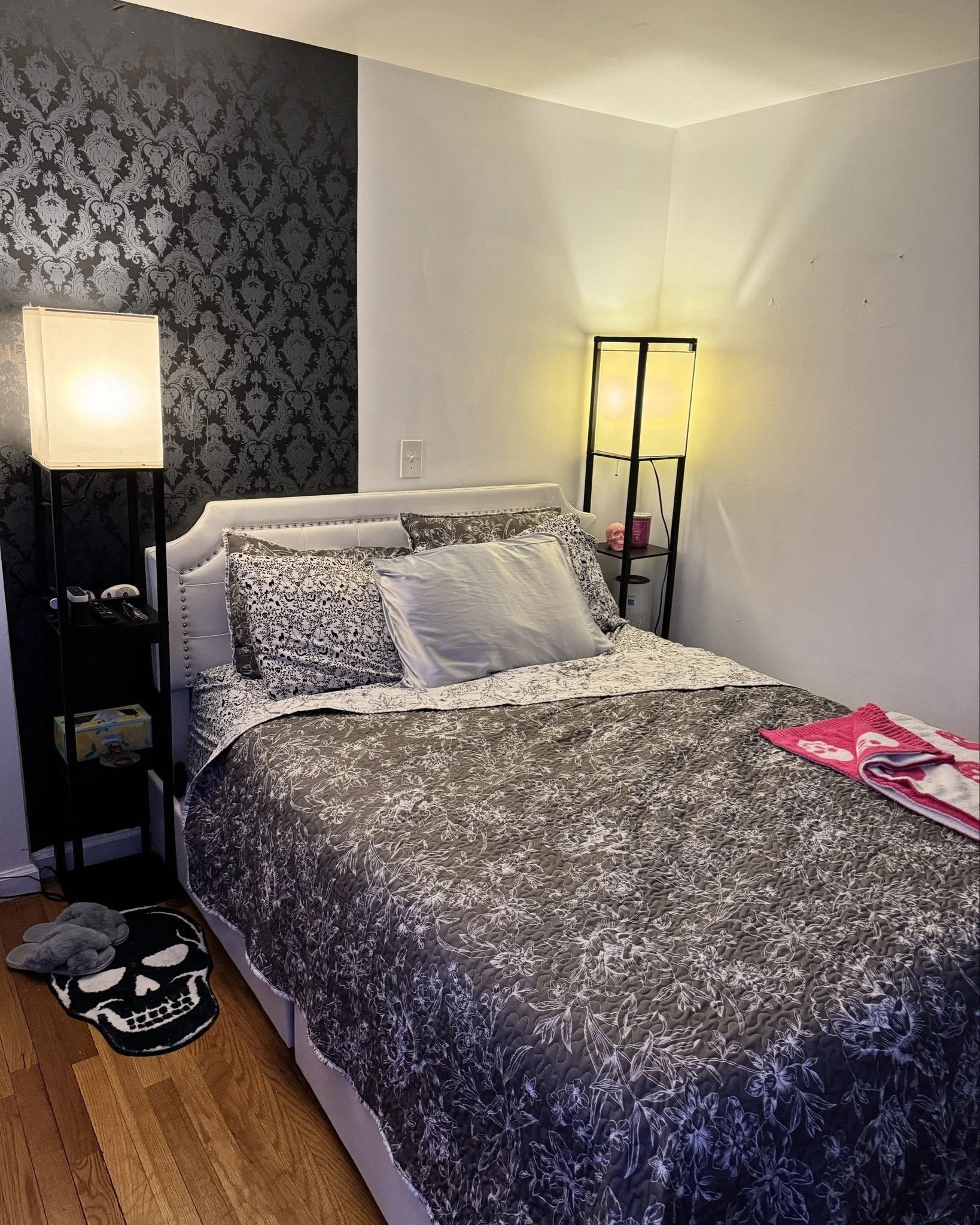 A bedroom with a bed covered in a floral patterned bedspread and pillows. There are two black standing lamps, one on each side of the bed. The wall behind the bed has a black and gray damask wallpaper on the left side and plain white on the right. A 