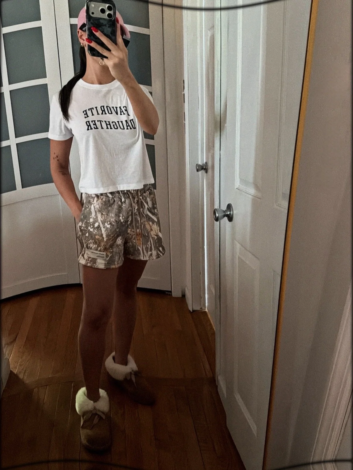 A woman takes a mirror selfie showing her outfit, which includes a white t-shirt with black text, camouflage shorts, and fuzzy slippers. She is in a room with wooden flooring, a door, and a wardrobe.
