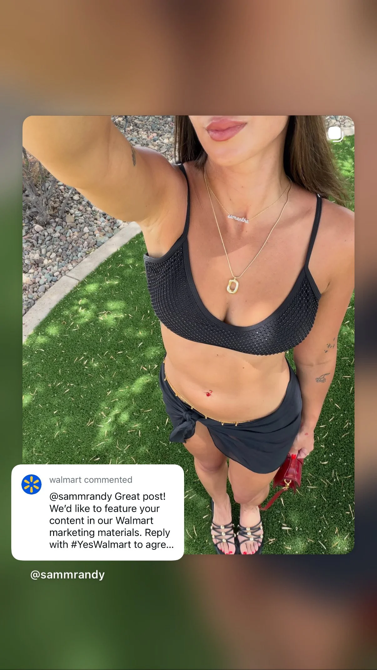 A woman is taking a selfie outdoors, wearing a black sports bra and black shorts. She is standing on a green lawn with a rocky area nearby. She has several necklaces, including one with the name 'Samantha', and visible tattoos on her arms and stomach