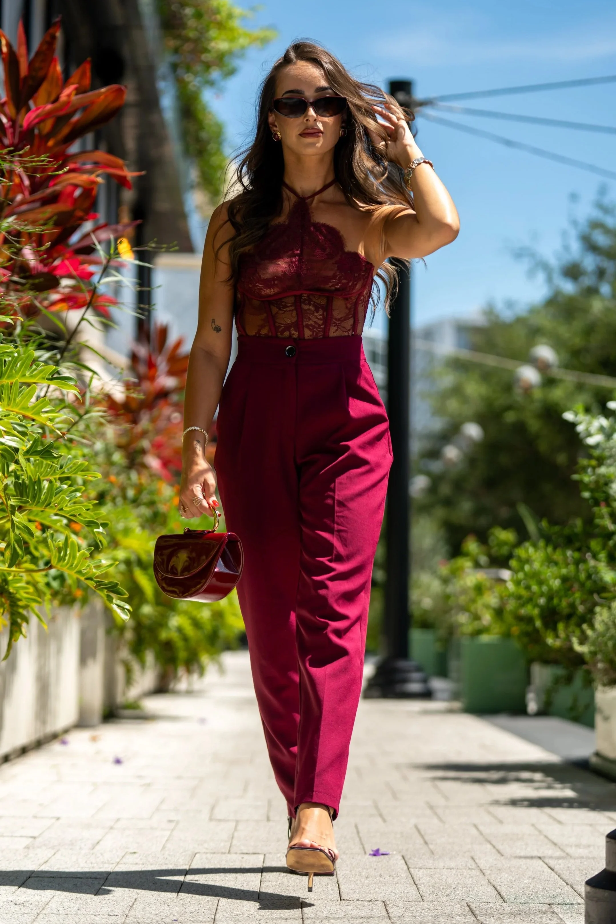 A woman wearing sunglasses, a burgundy lace top, and matching pants, walking on a sidewalk with plants on her sides and a clear blue sky overhead.